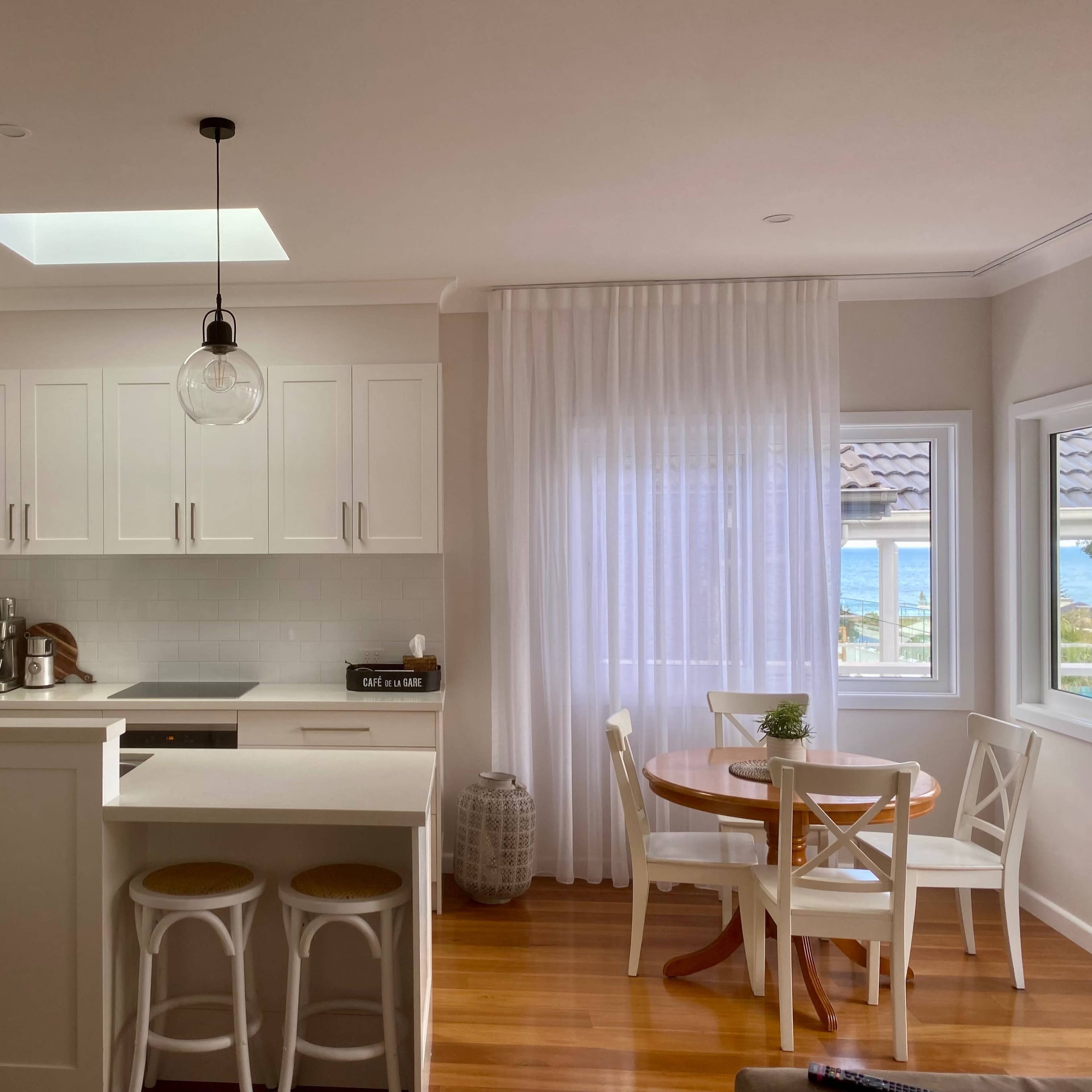 A bright kitchen features white cabinetry, pendant lights, and a dining area with a round table and views of the sea through large windows.