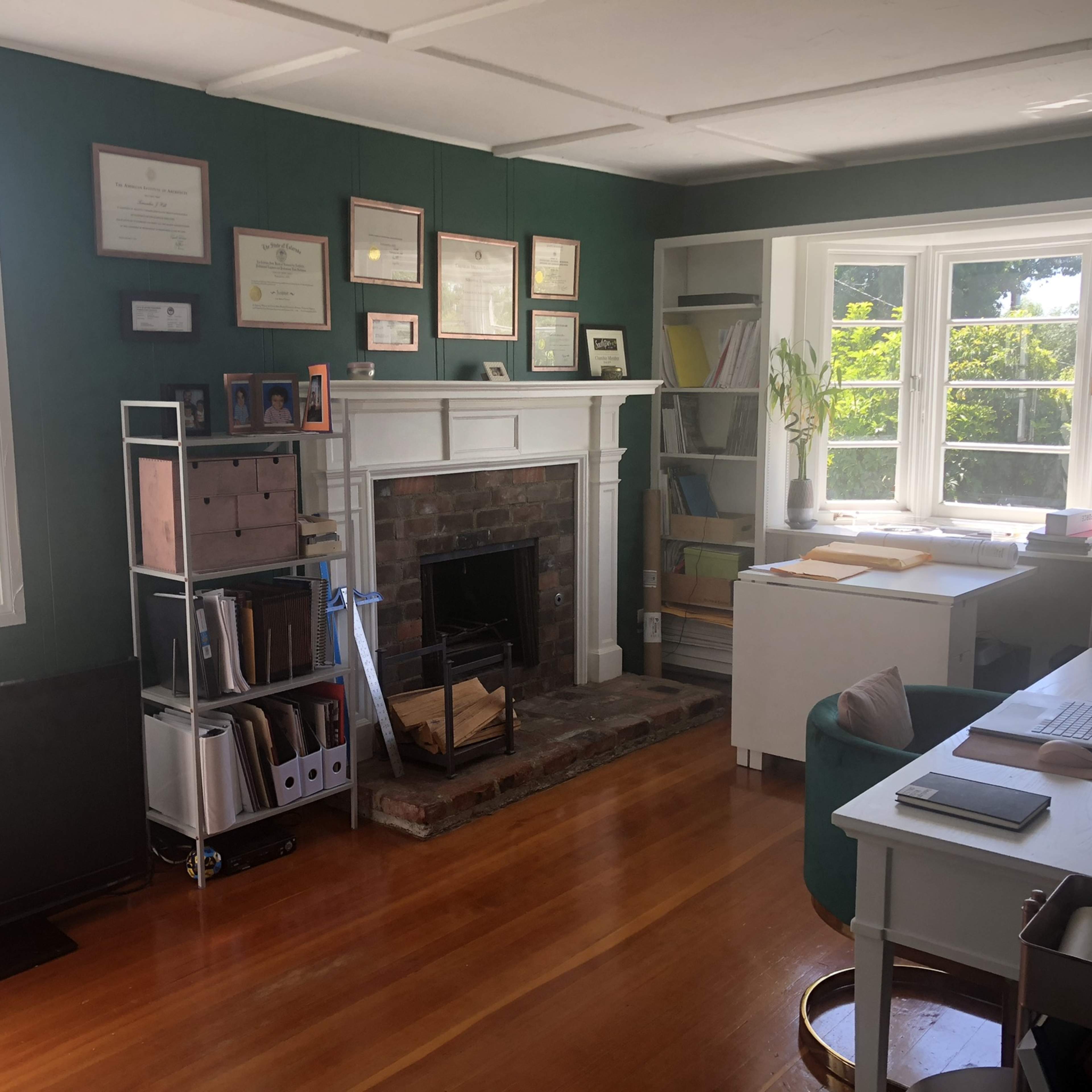 A home office with a fireplace, desks, shelves filled with books and documents, and large windows letting in natural light.