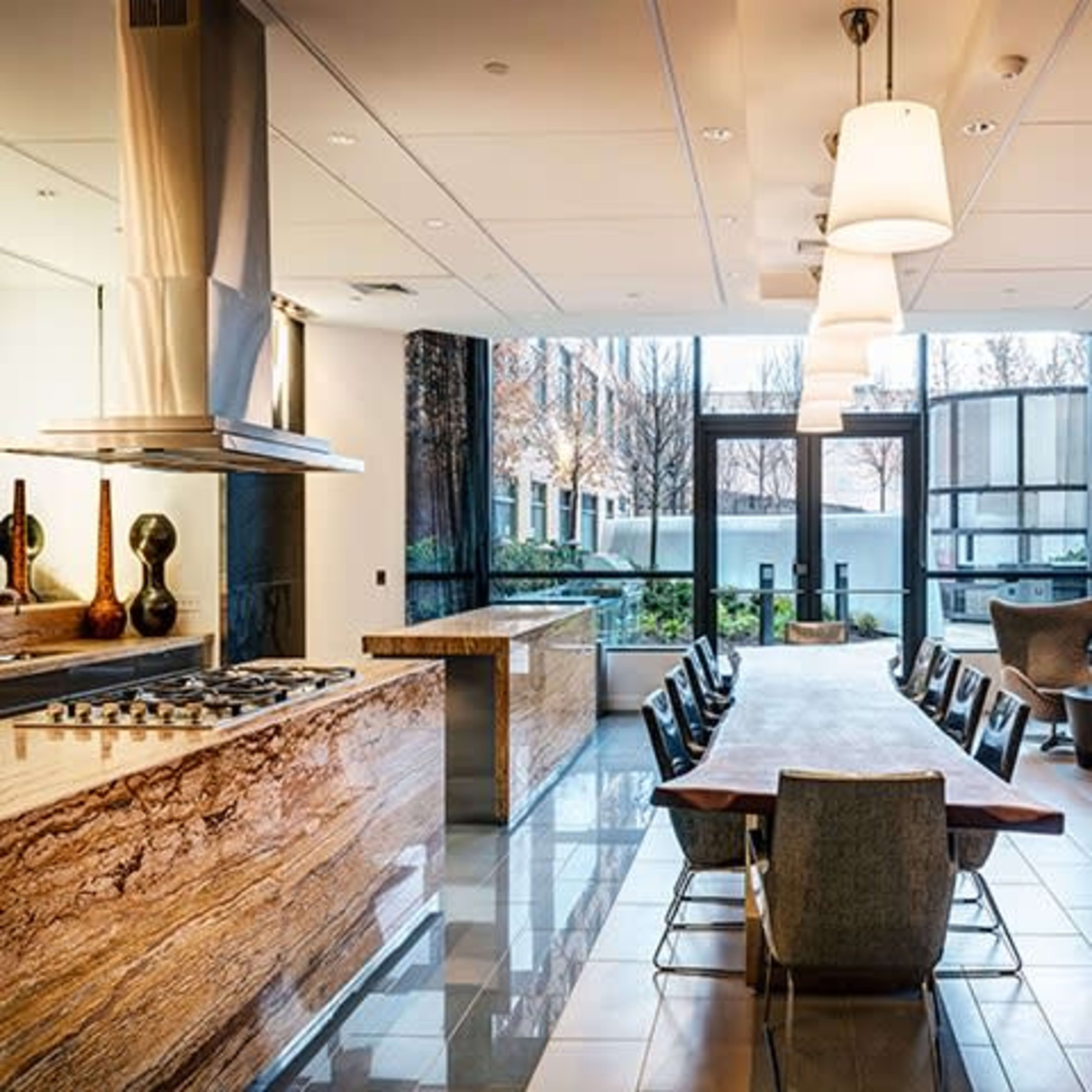 A modern kitchen and dining area features a large wooden table surrounded by chairs, with a sleek kitchen setup, and large windows offering views of the outdoors.