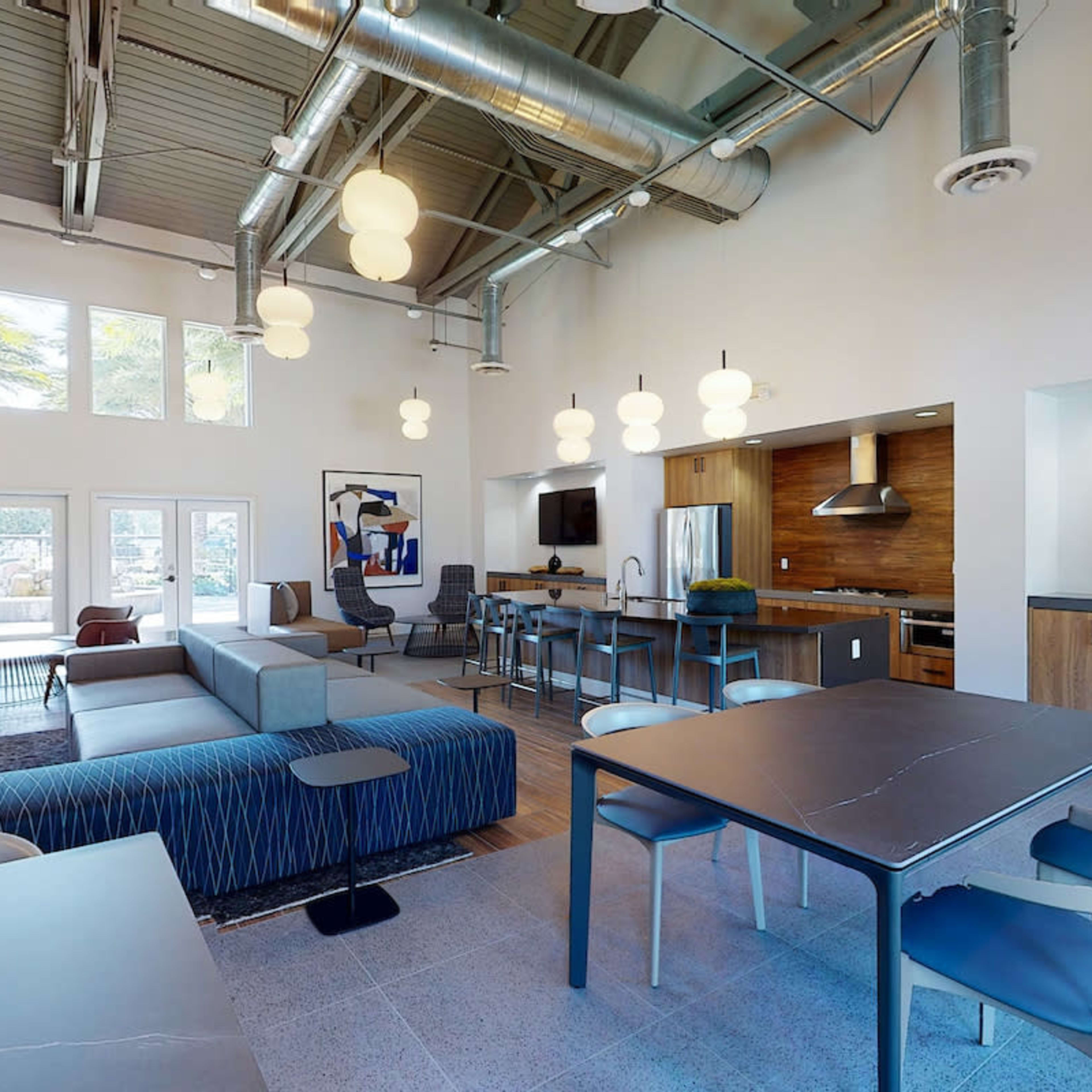 A modern communal lounge area featuring a seating arrangement, a kitchen, and dining tables under pendant lighting.