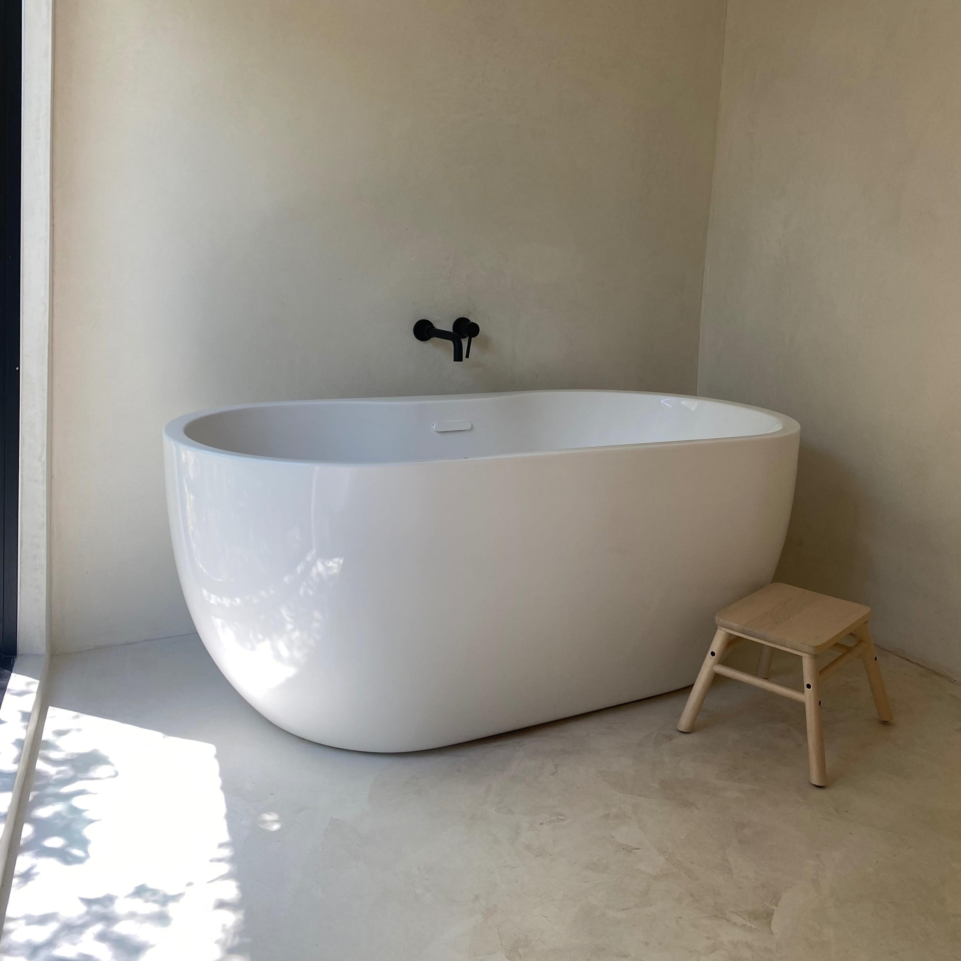 A modern, freestanding white bathtub beside a small wooden stool in a minimally designed bathroom with light-colored walls.