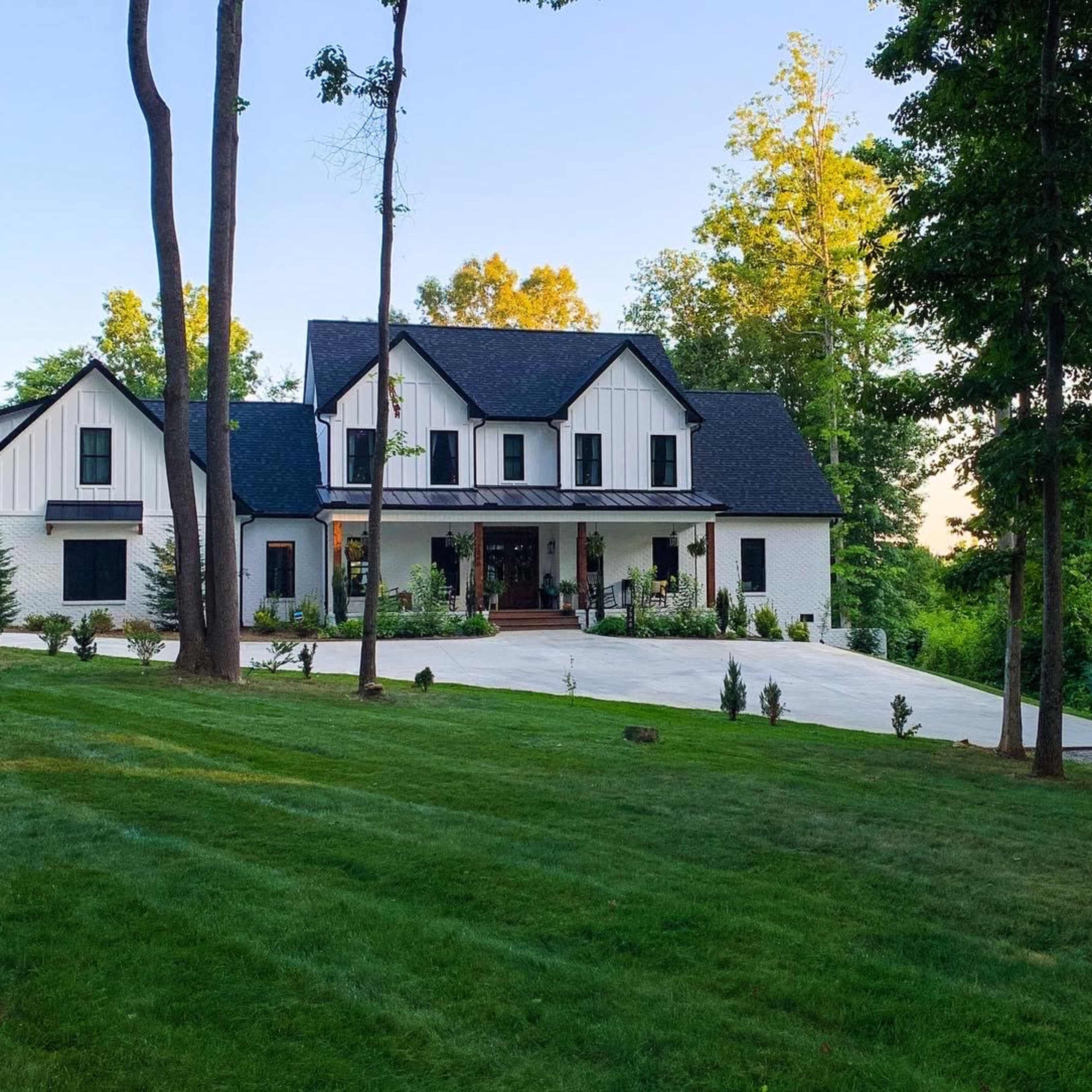 A modern two-story house with a black roof and white facade is situated on a green lawn surrounded by trees.
