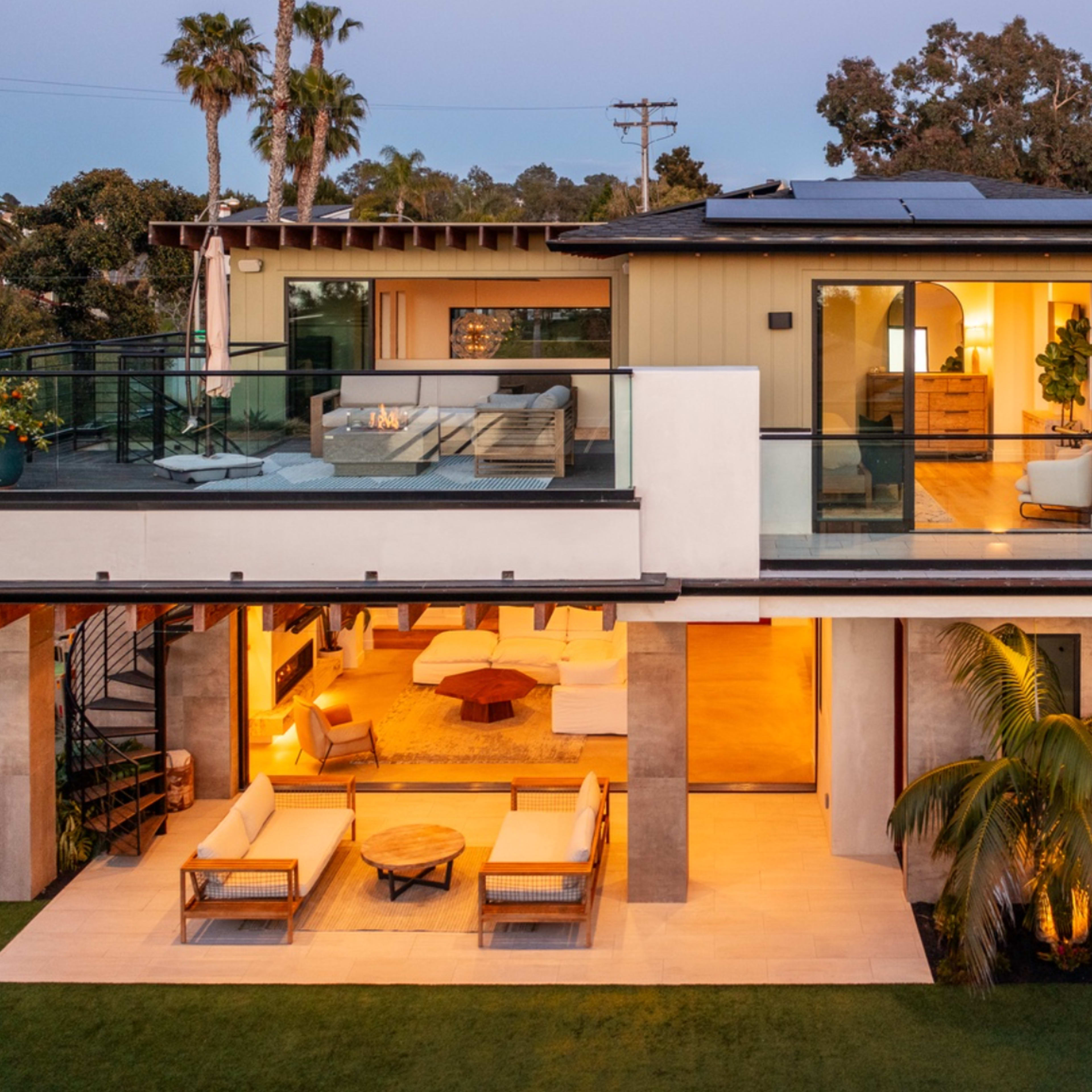 A modern two-story house features large glass windows, a spacious balcony with seating, and a well-lit interior visible from the patio.