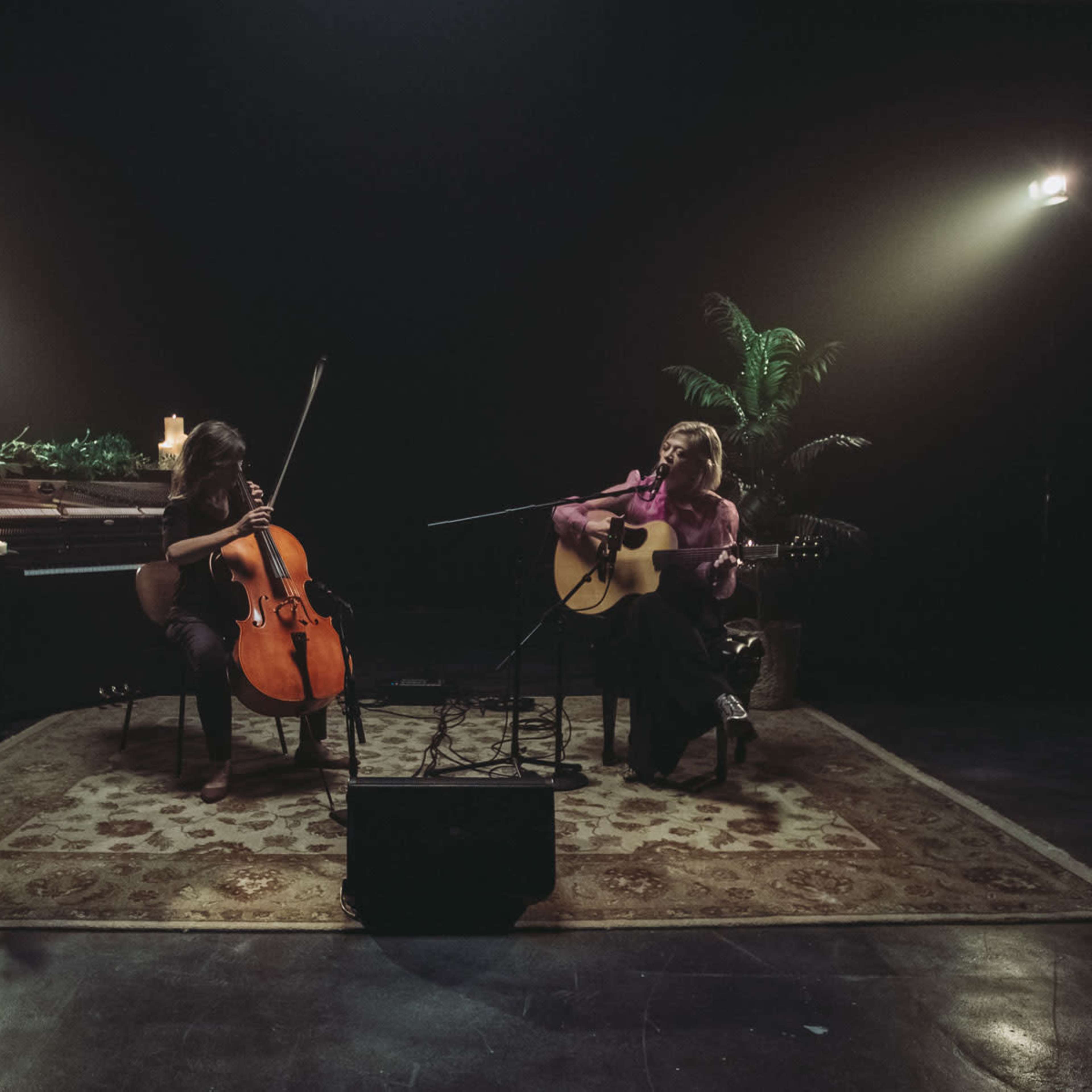Two musicians are performing together, one playing a cello and the other an acoustic guitar, in a dimly lit room with candles and plants.