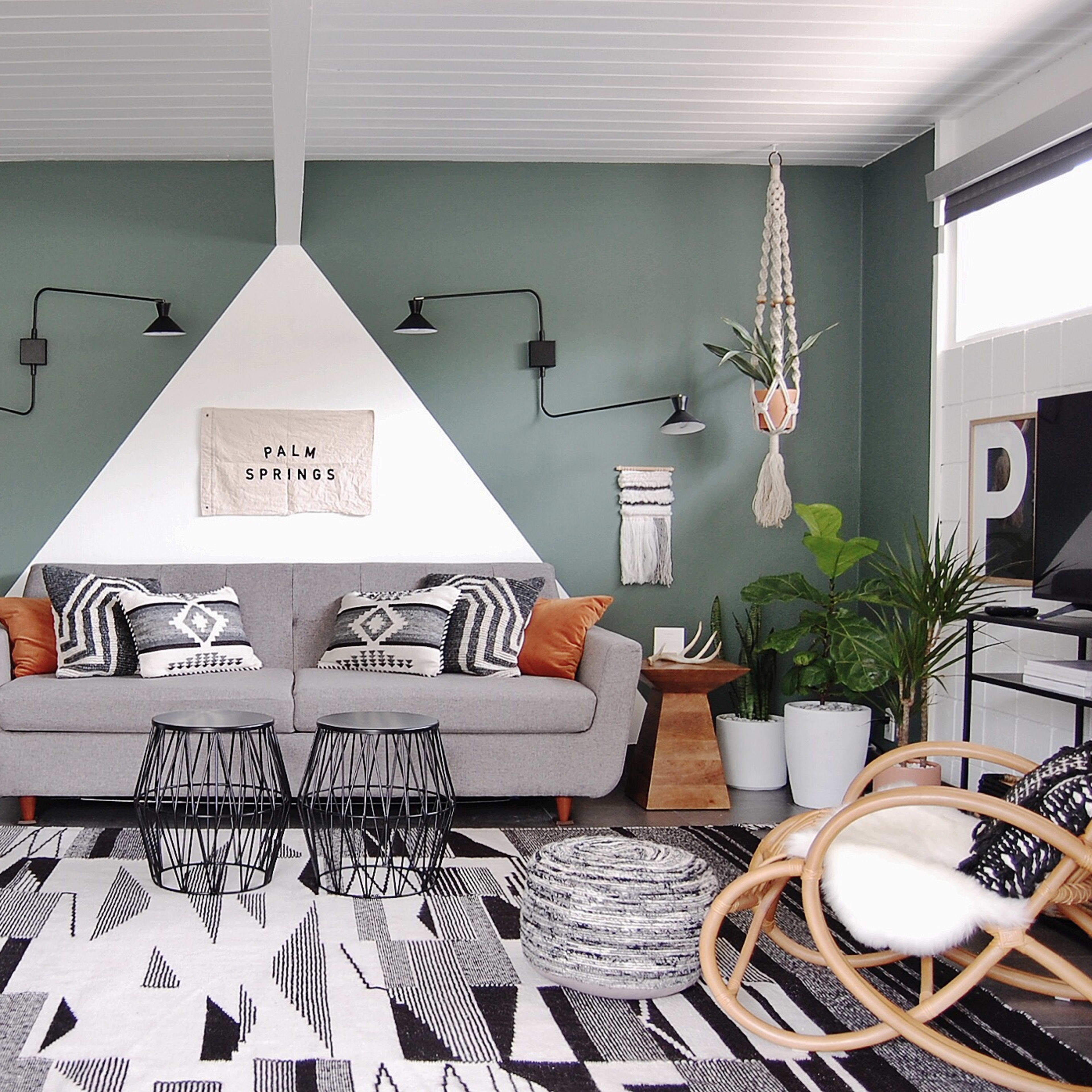 A modern living room features a gray sofa with patterned pillows, two geometric side tables, and an area rug with black and white designs, set against a green accent wall.