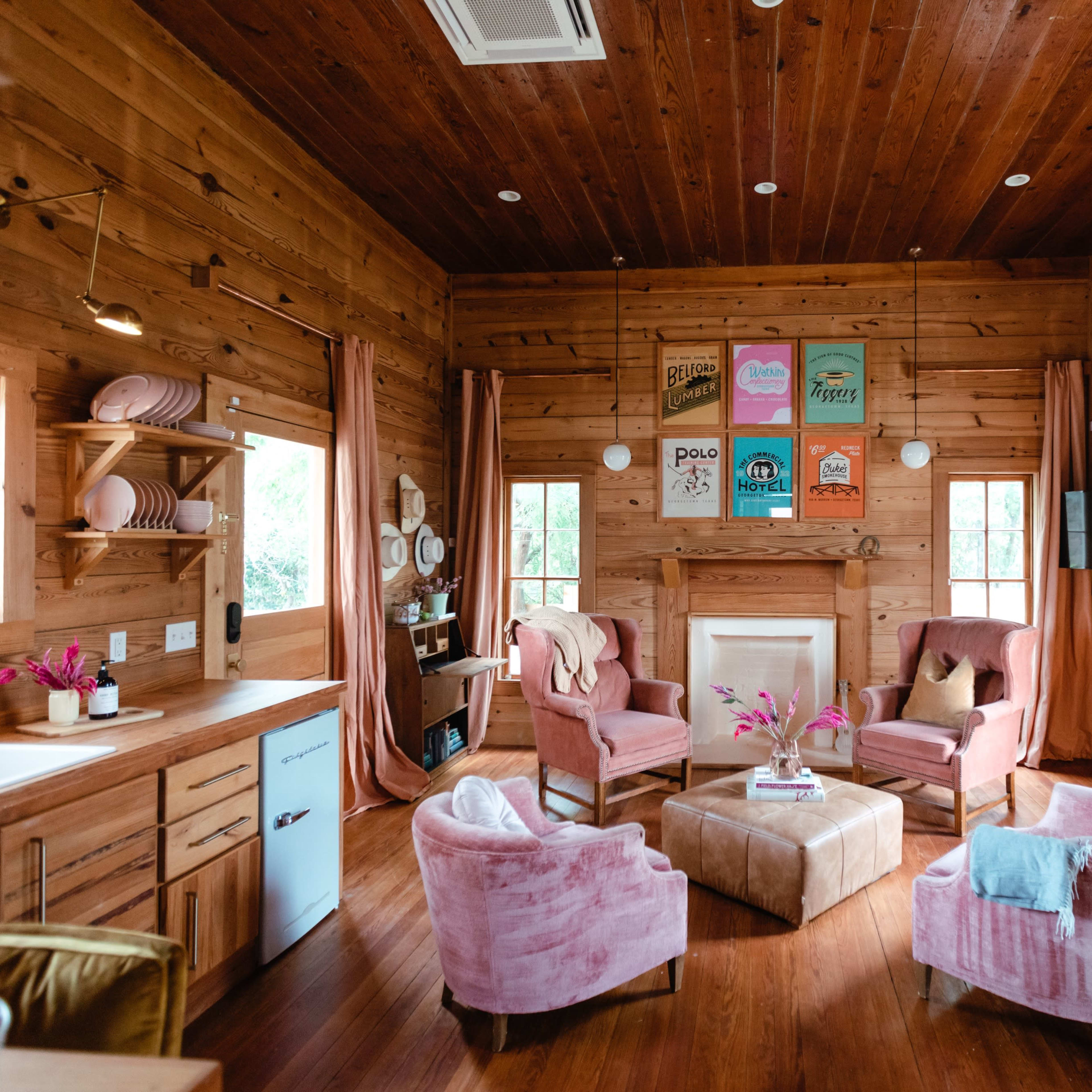 A cozy wooden cabin living room with pink upholstered chairs, a coffee table, and colorful artwork on the walls.