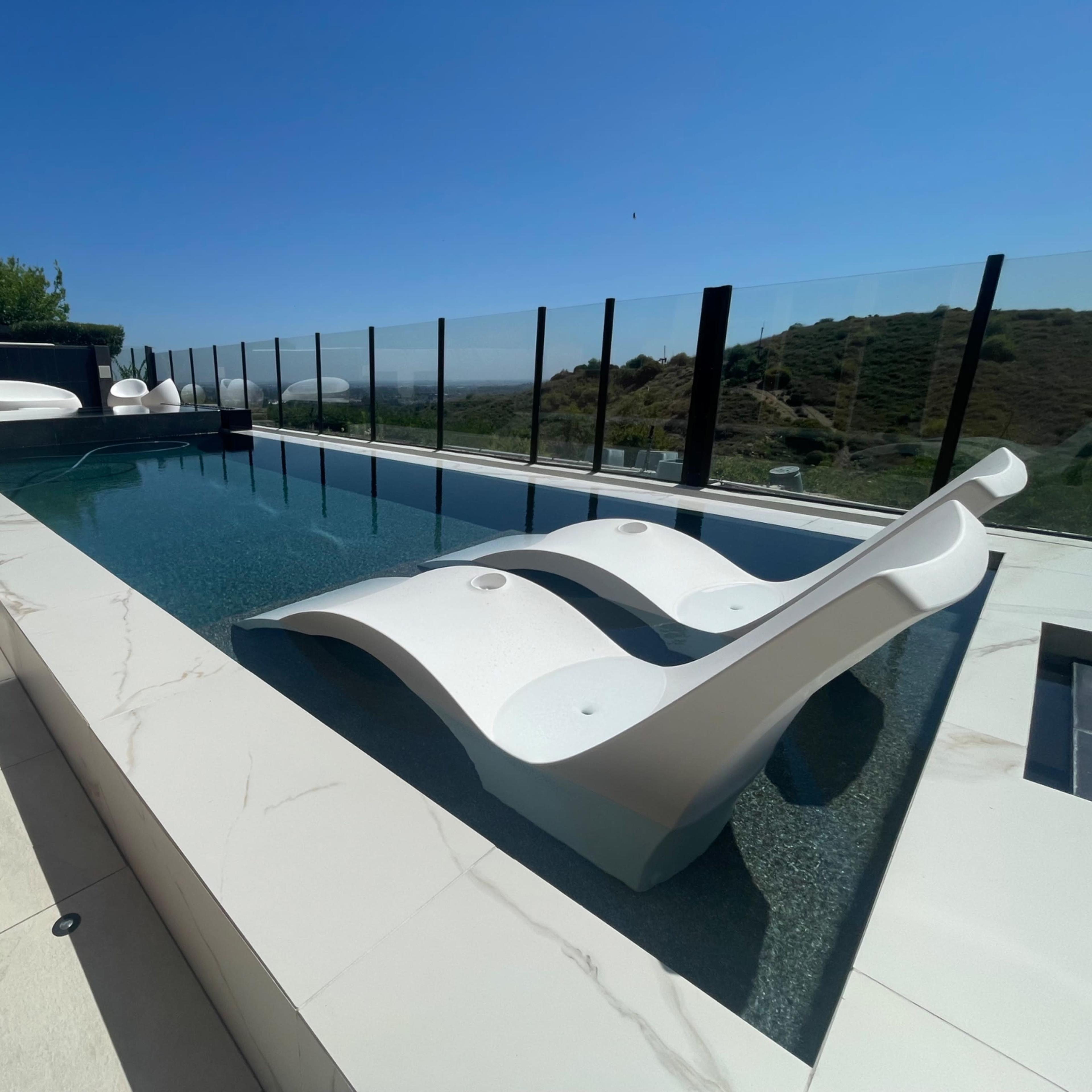 A modern infinity pool with white loungers and a clear glass railing overlooking a hillside.