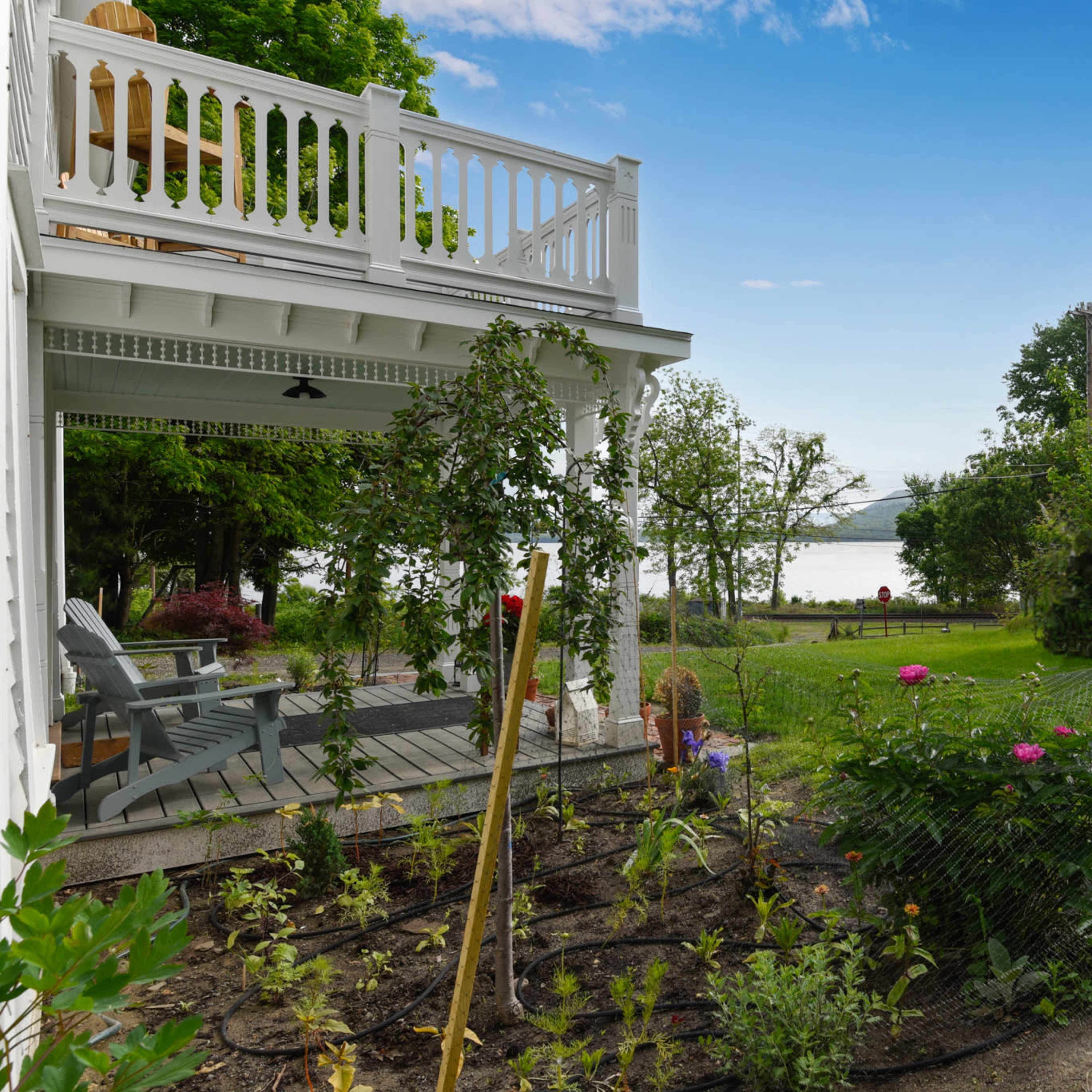 A house with a porch and a garden overlooks a lake, surrounded by trees and a grassy yard.