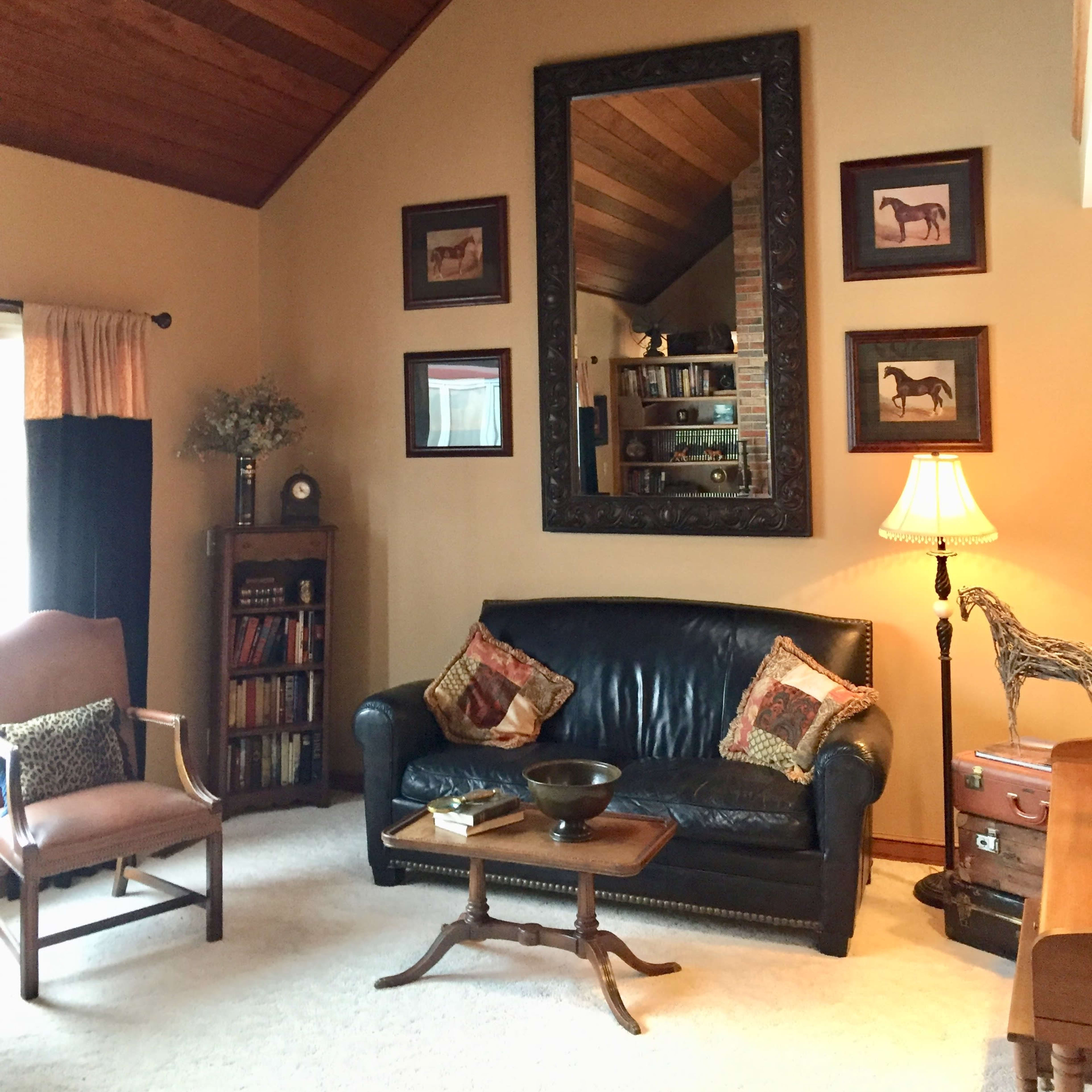 A living room with a leather sofa, a wooden ceiling, framed horse prints on the walls, and a lamp next to a chair.