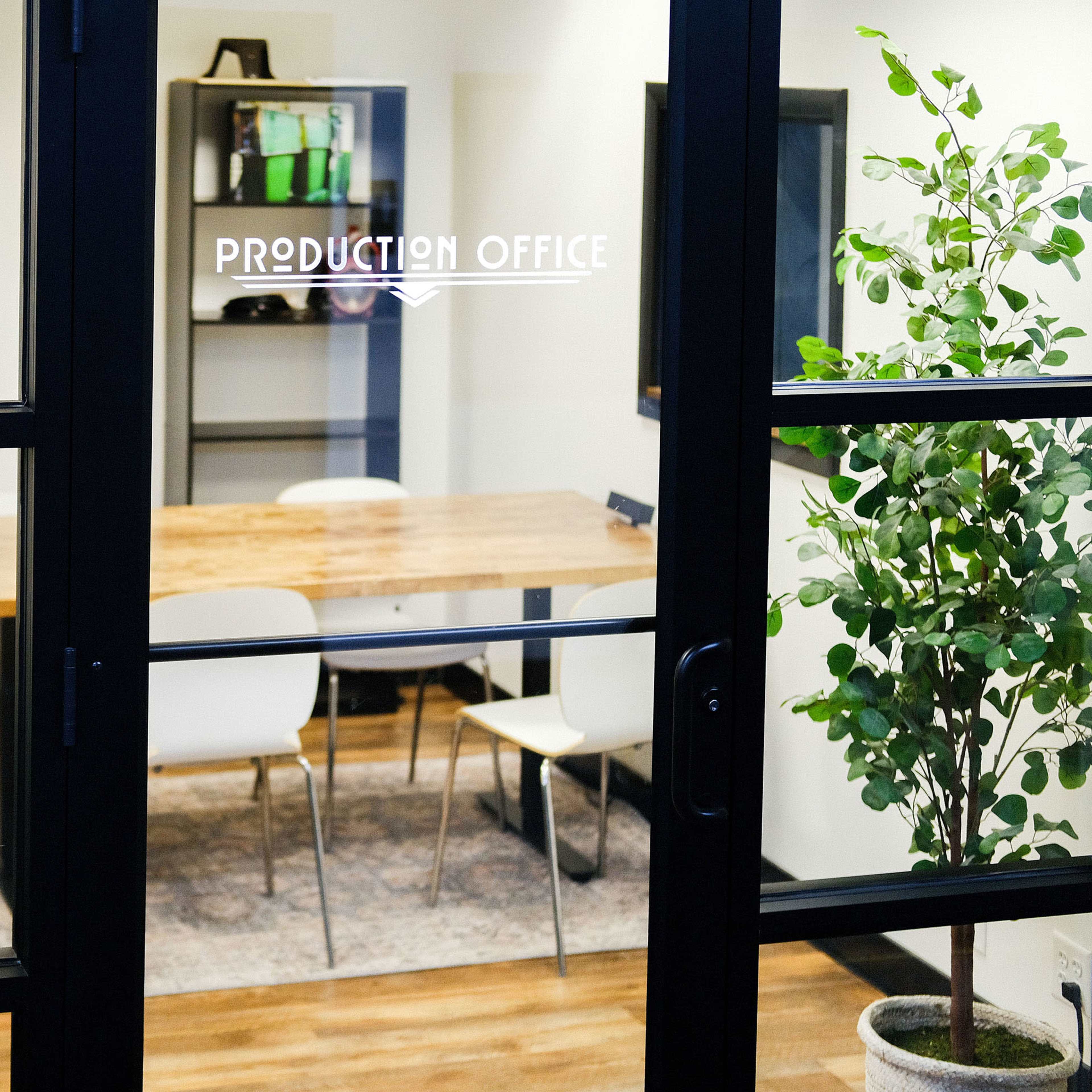A production office features a wooden table surrounded by white chairs, with a potted plant visible through the glass doors.