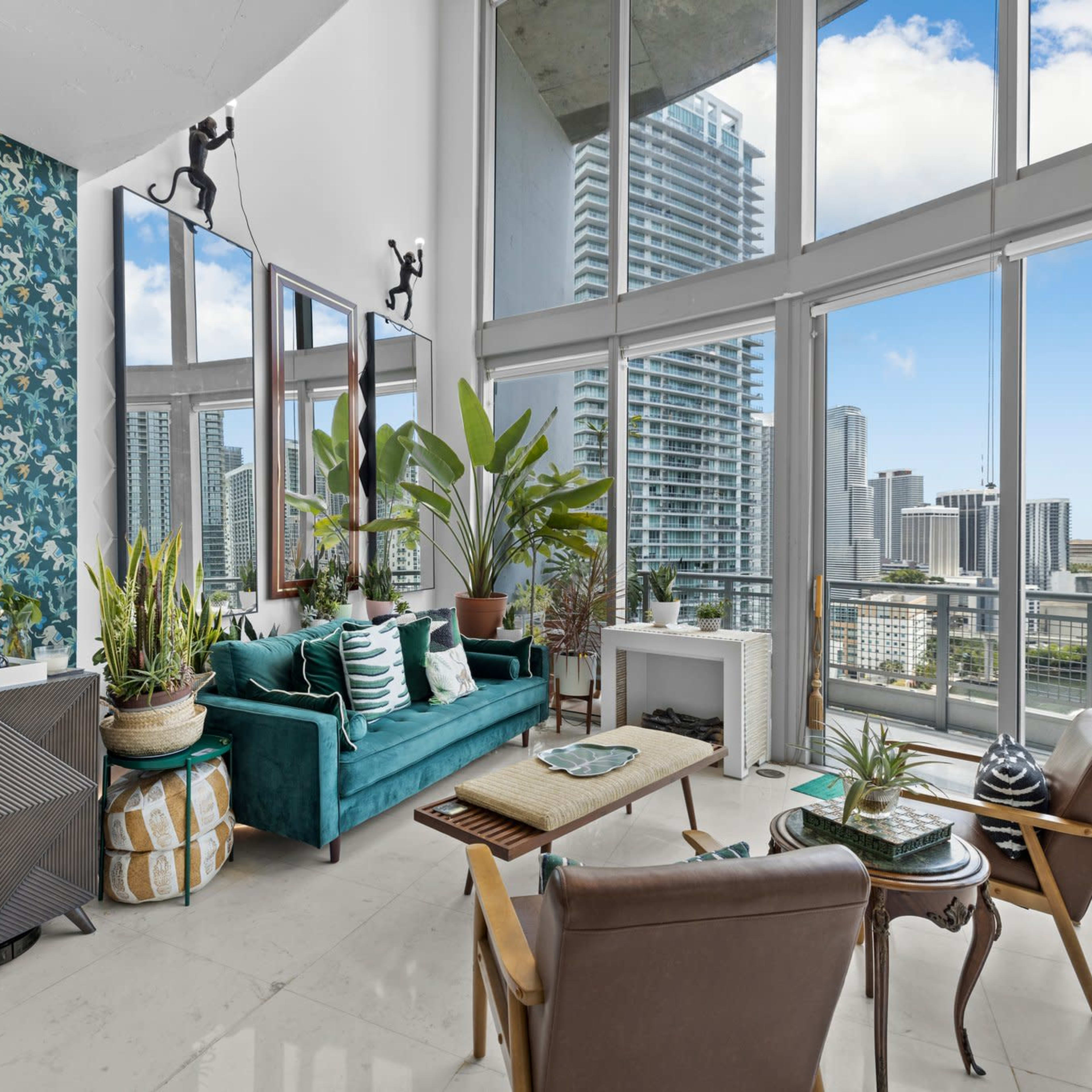 A modern living room features a teal sofa, a round mirror, and large windows showing a view of city skyscrapers, along with various plants and stylish furniture.