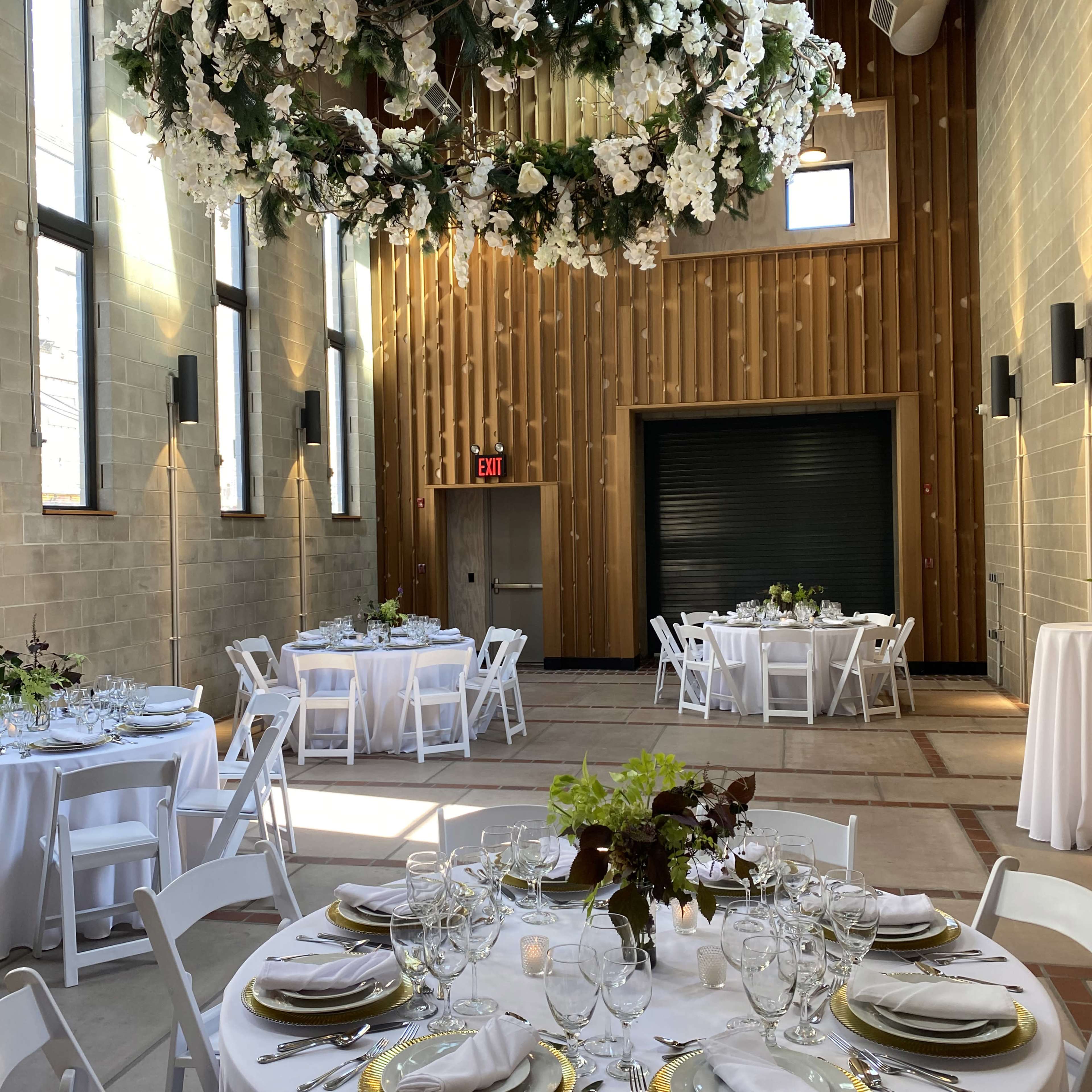 A beautifully arranged indoor event space with white tables set for dining, surrounded by wooden walls and a floral ceiling installation.