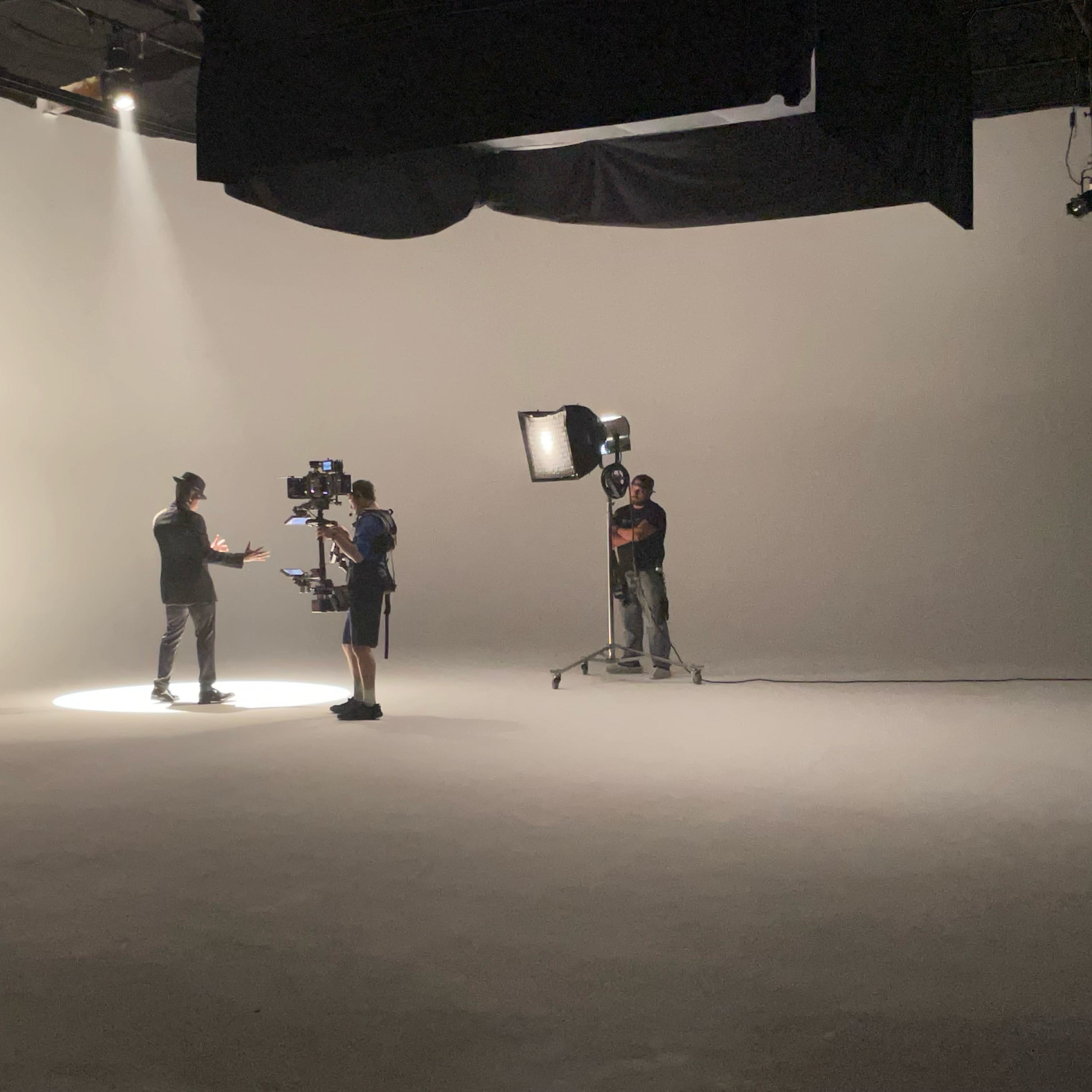 A film crew is set up in a studio, with one person speaking under a spotlight while a camera operator films them.