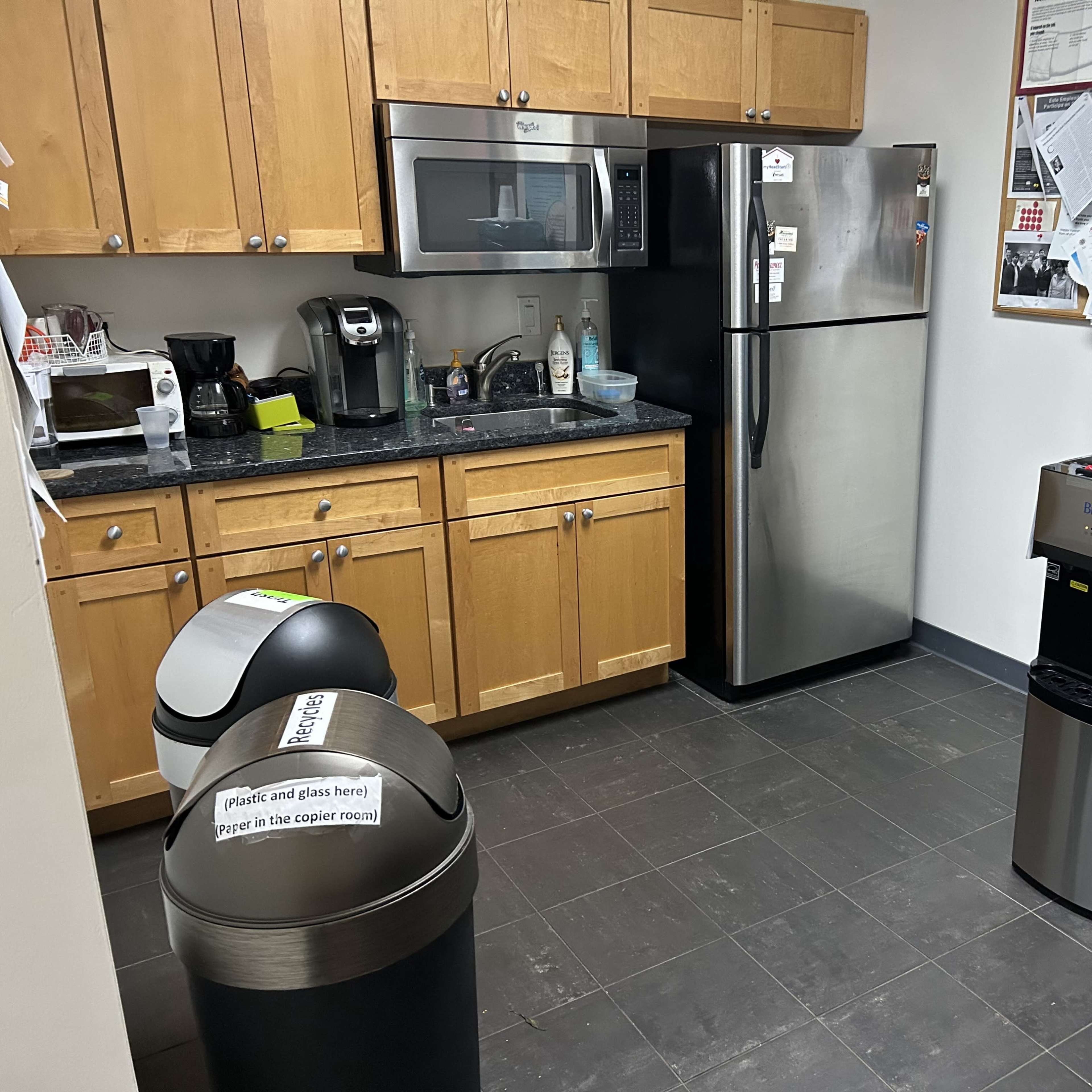 A kitchen area with wooden cabinets, a microwave, a stainless steel refrigerator, and a water cooler next to two trash bins.