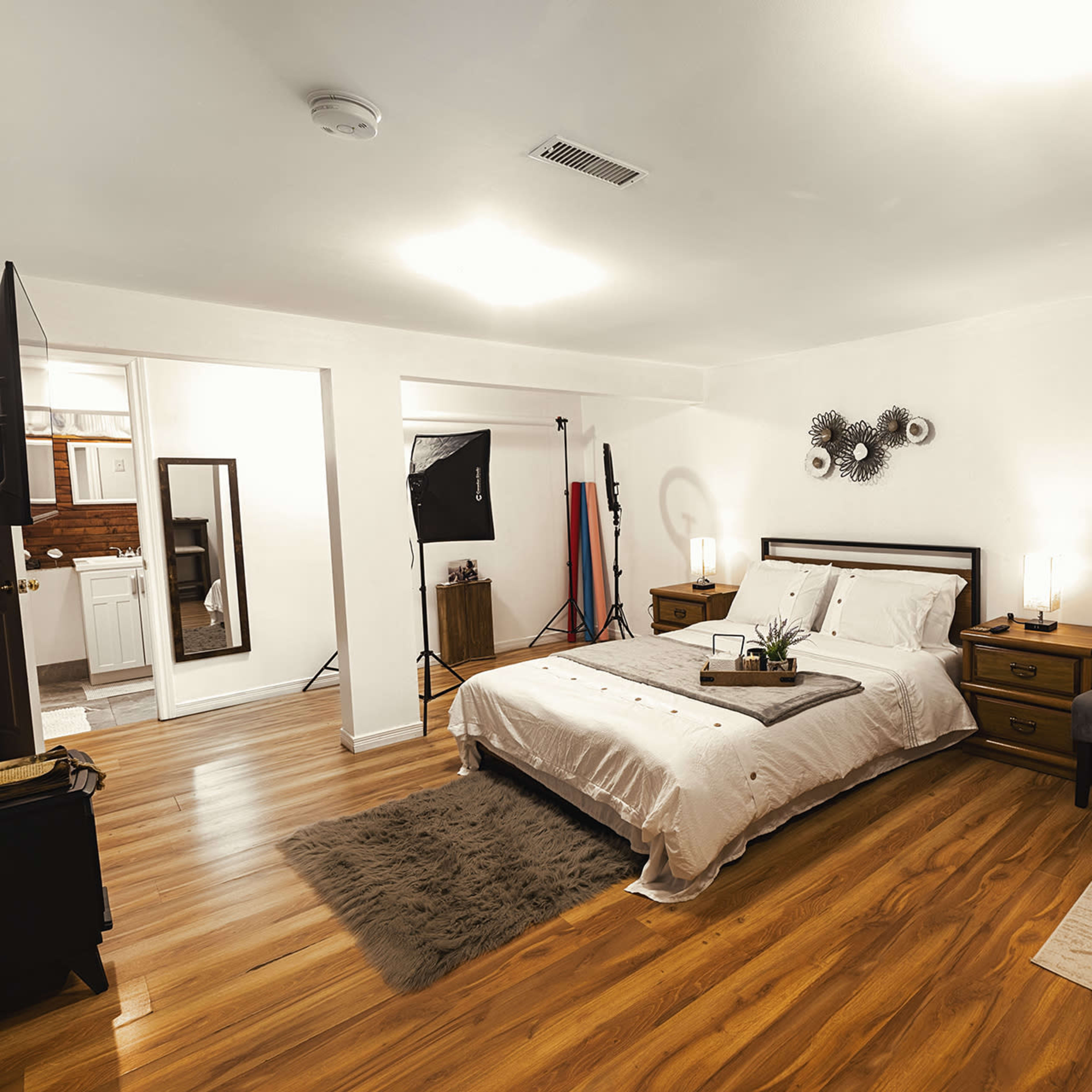 A well-lit bedroom featuring a bed with white linens, two nightstands, a chair, and a small table, all set on a hardwood floor.