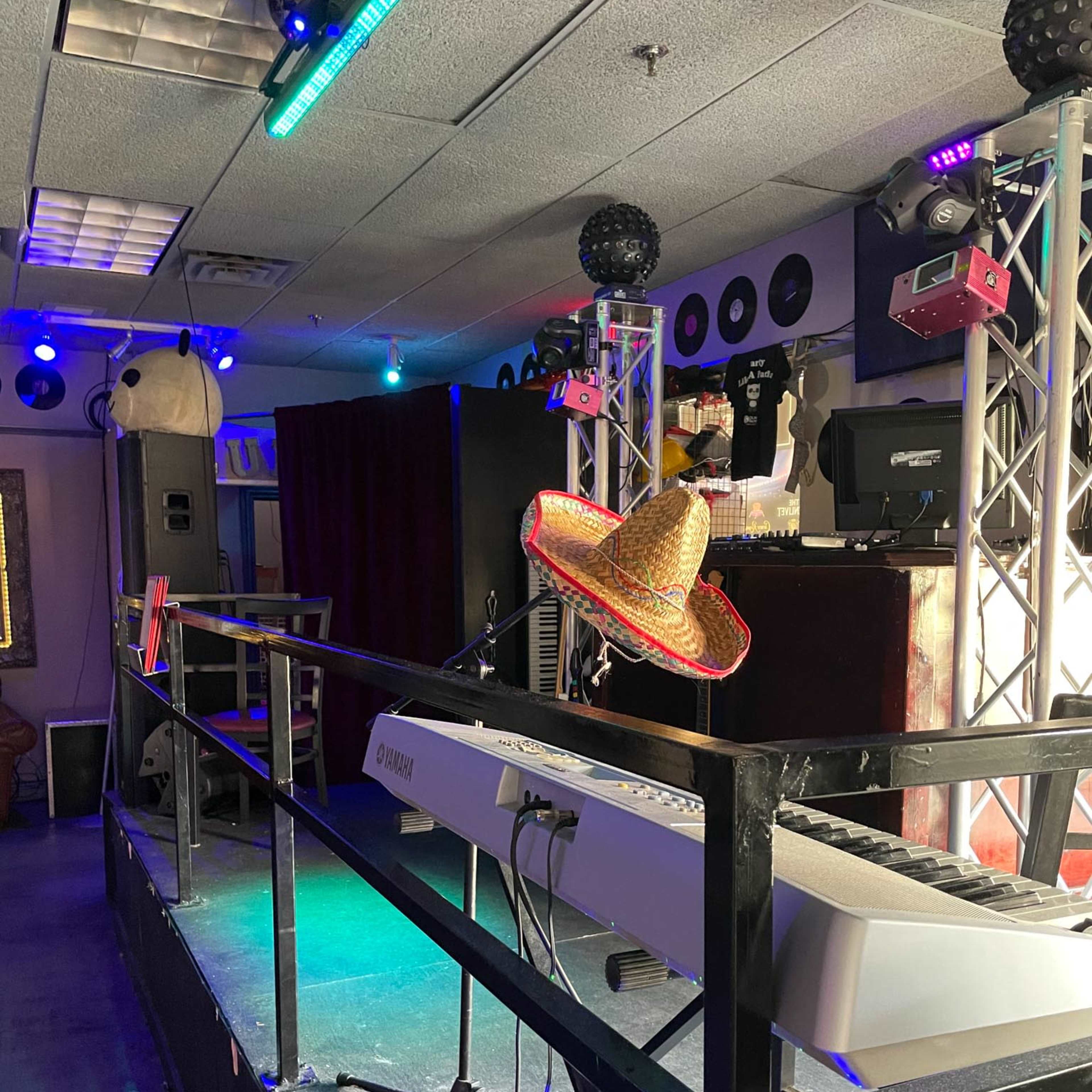 A small stage in a music venue with a keyboard, a sombrero placed on it, and colorful lights in the background.
