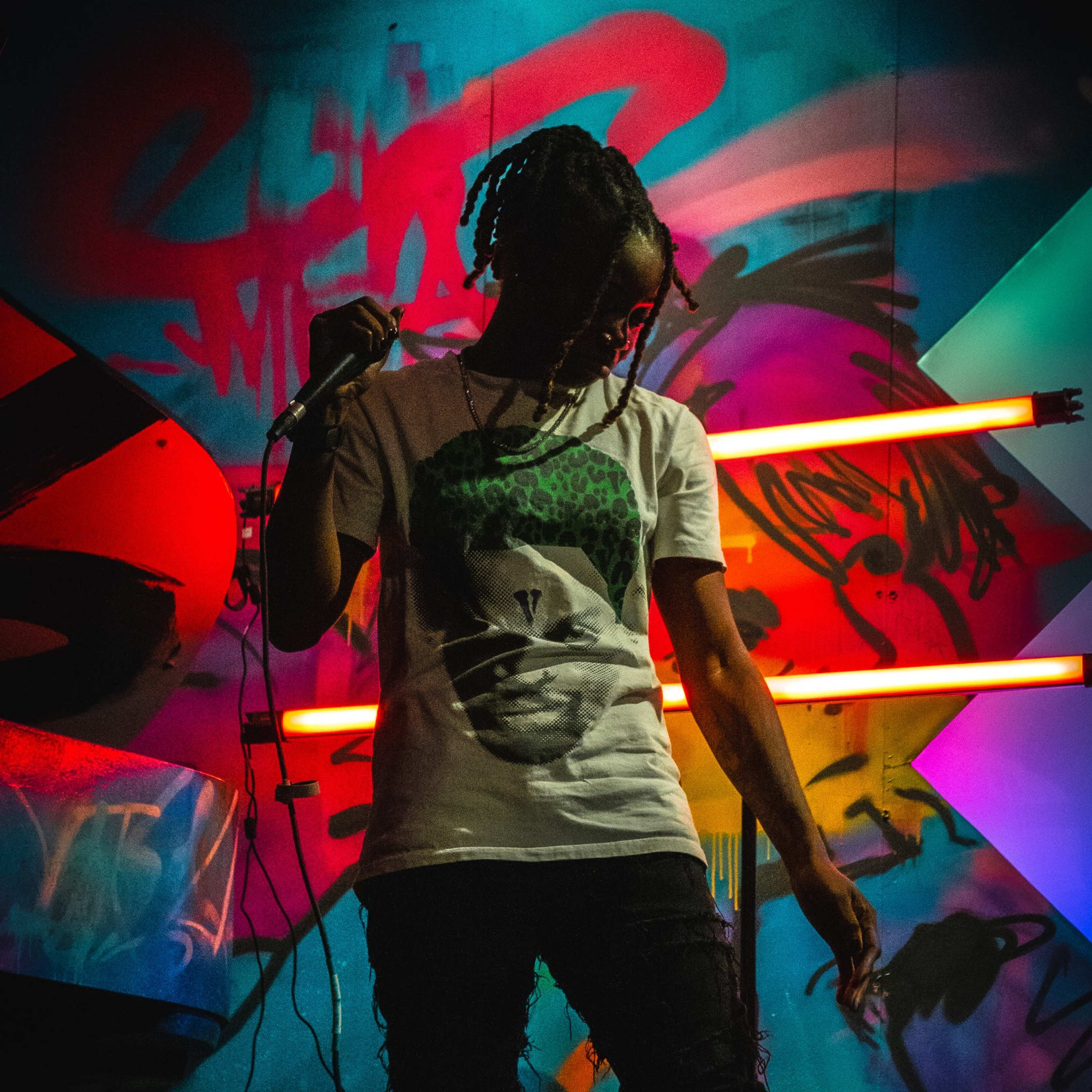 A person stands on stage holding a microphone in front of a colorful, graffiti-covered backdrop with neon lights.