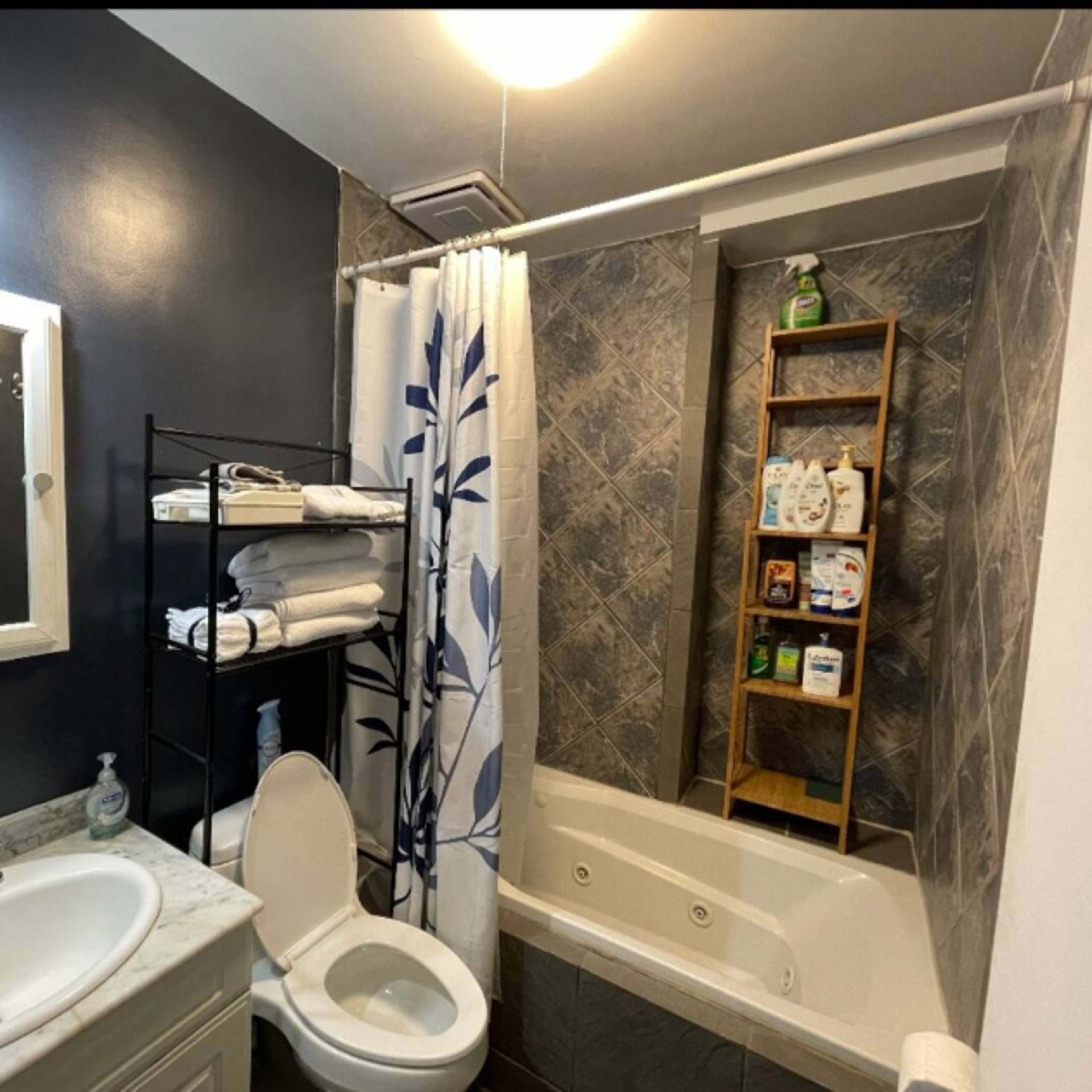 A compact bathroom featuring a tub, a toilet, a mirrored cabinet, and a shelving unit with neatly arranged toiletries.
