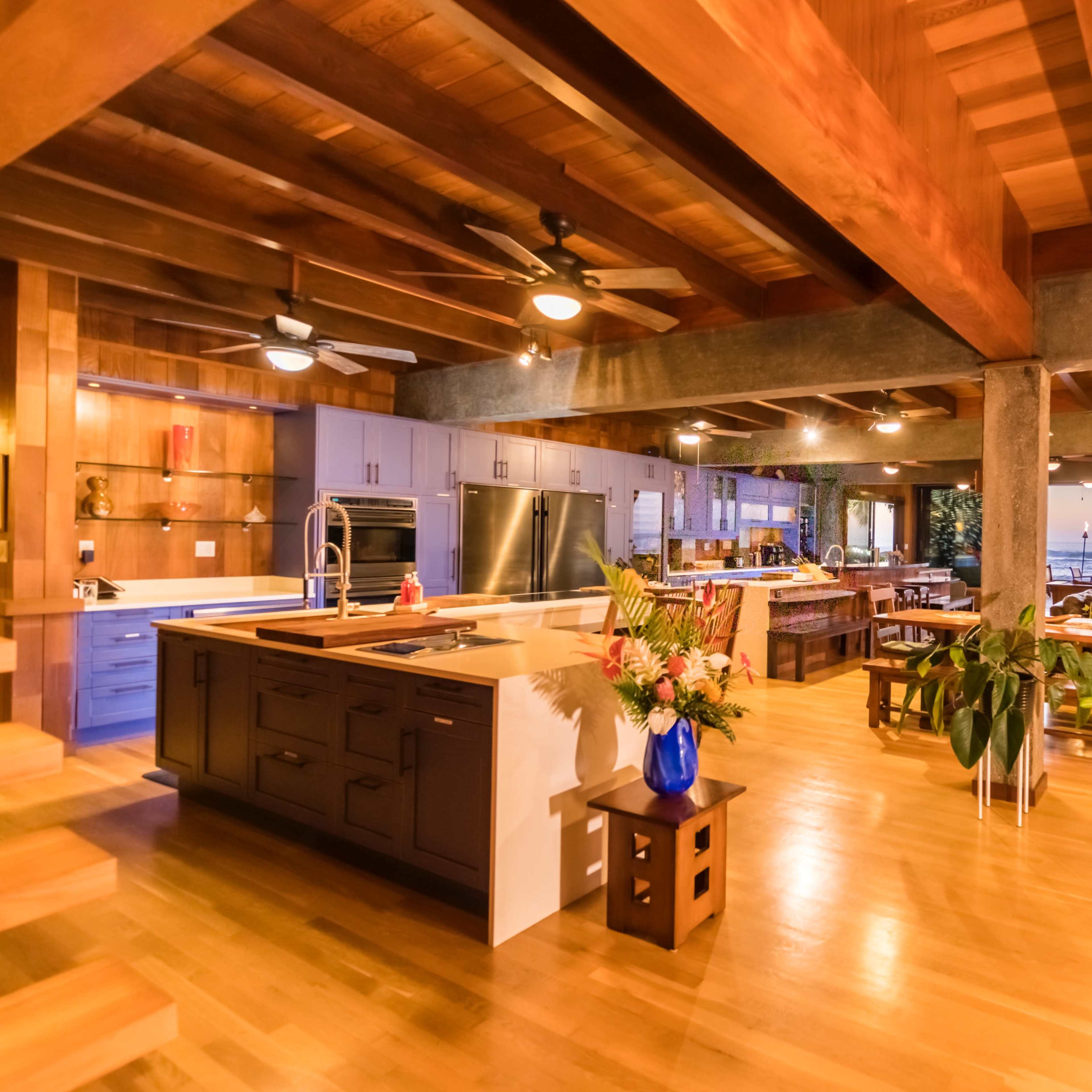 A spacious kitchen and dining area with wooden beams, large windows, and views of the ocean at sunset.