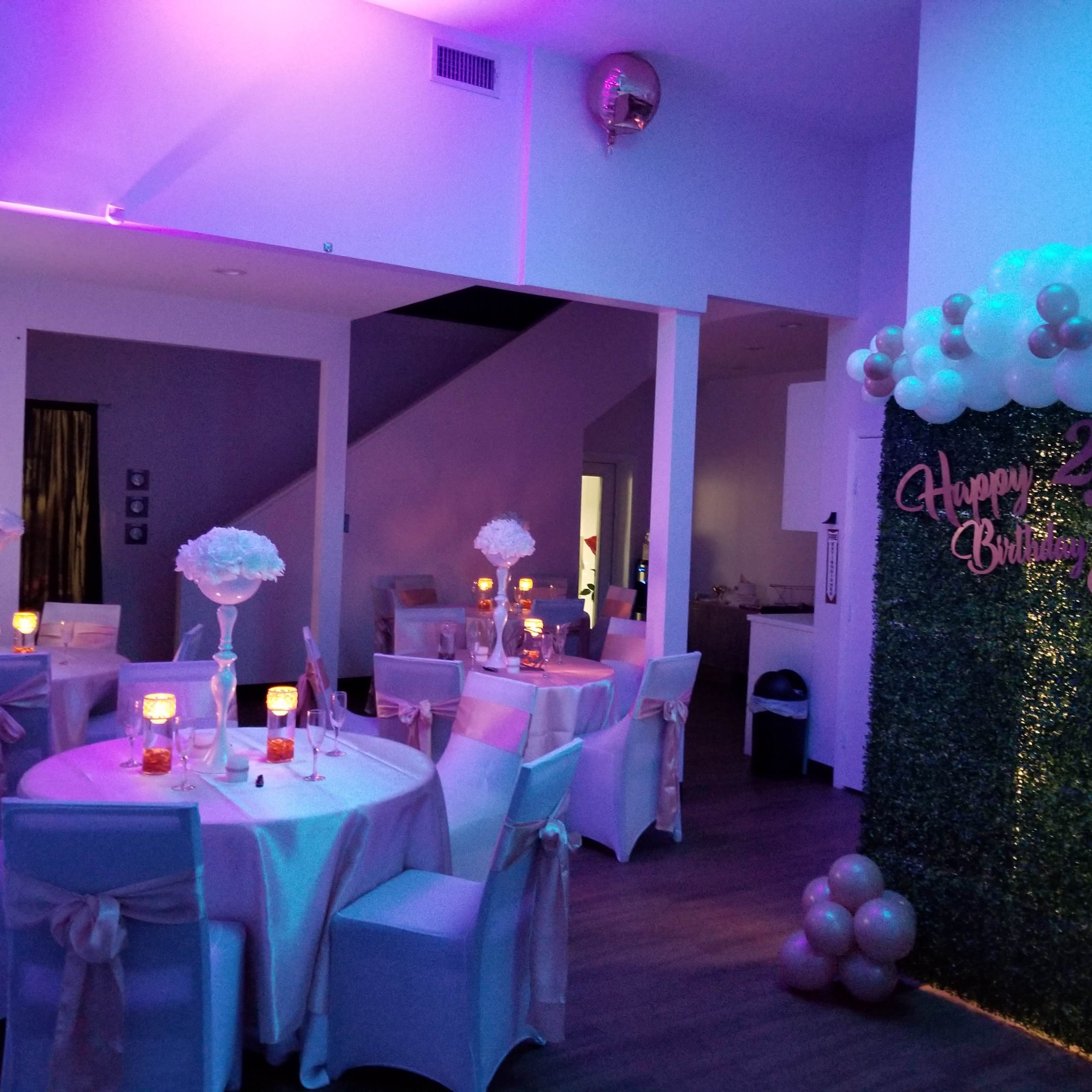 A decorated room set up for a birthday celebration, featuring tables with light-colored tablecloths, floral centerpieces, and a wall adorned with balloons and a birthday message.