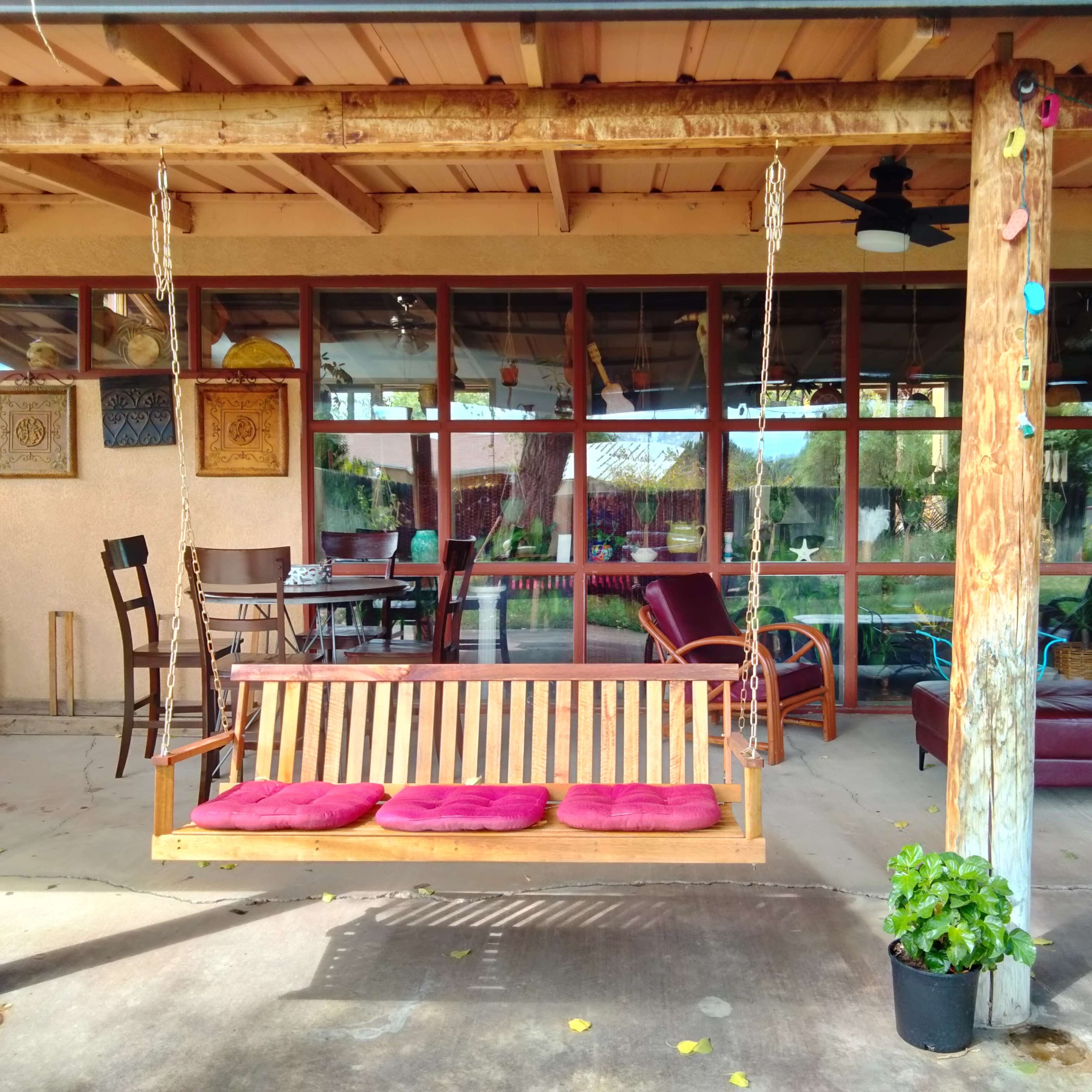 A porch with a wooden swing, surrounded by chairs and a table, and large windows overlooking a garden area.
