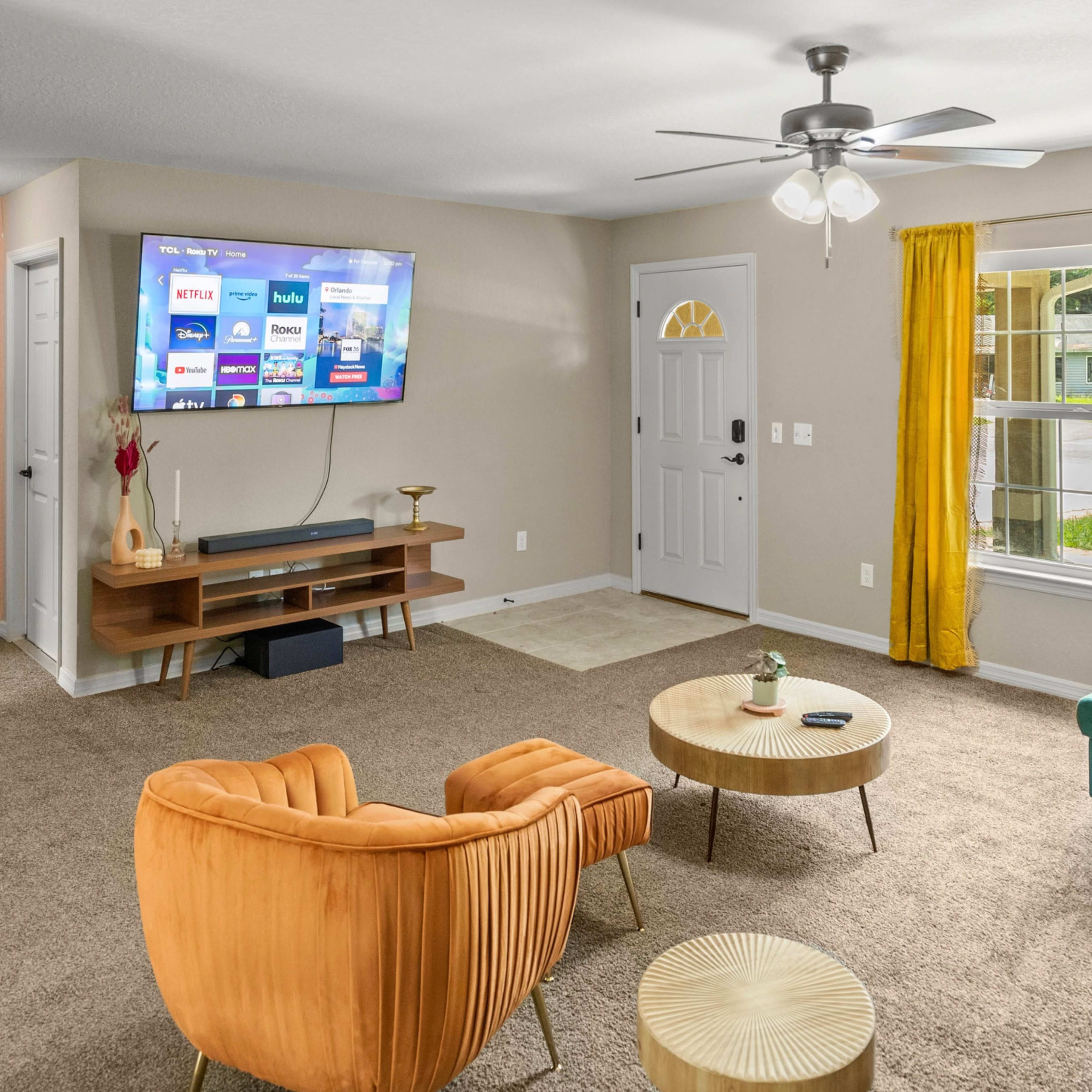 The living room features a television mounted on the wall, a green sofa, two orange chairs, and a large window with yellow curtains.