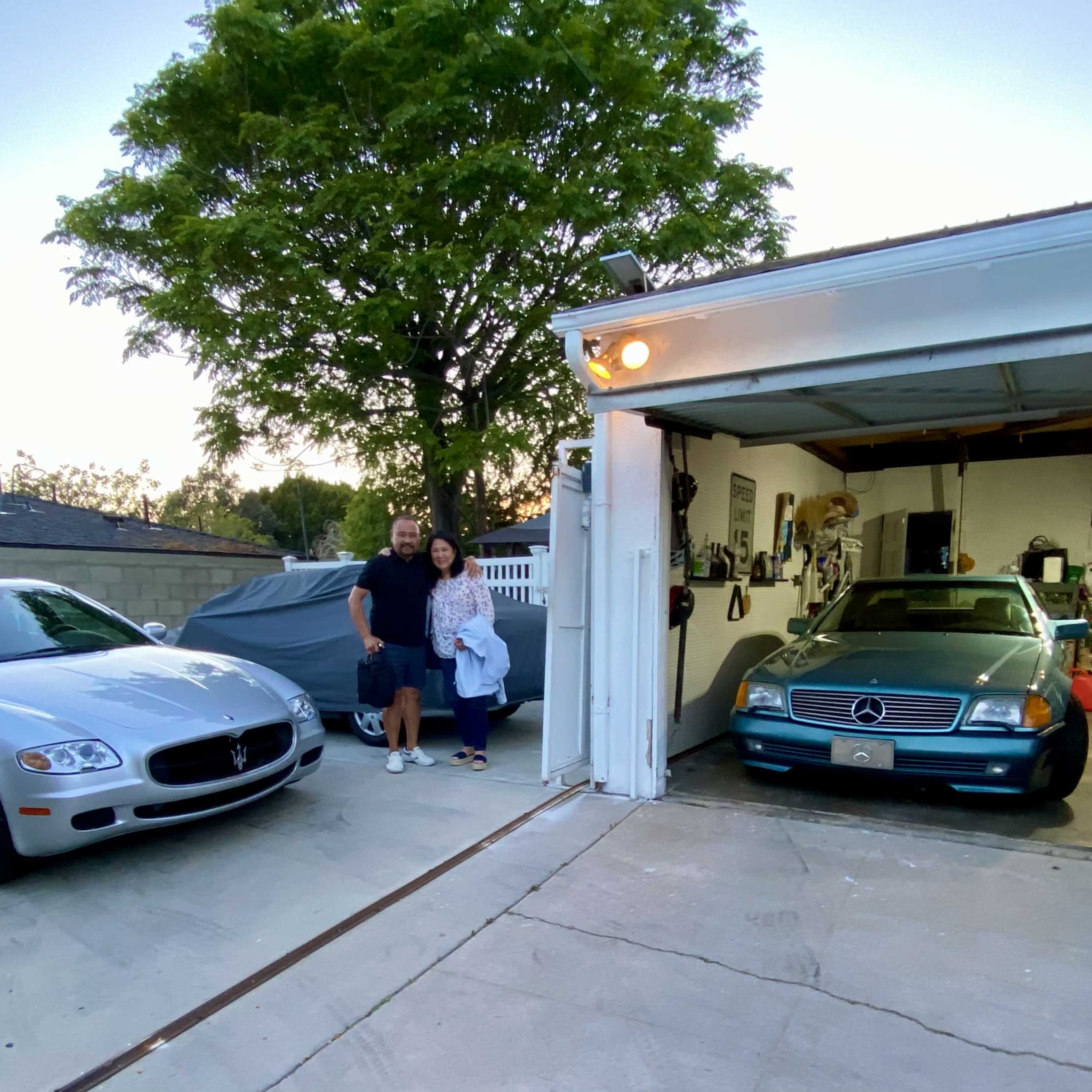 A couple stands in front of a garage beside two parked cars, one a silver Maserati and the other a blue Mercedes-Benz, with additional vehicles visible inside the garage.