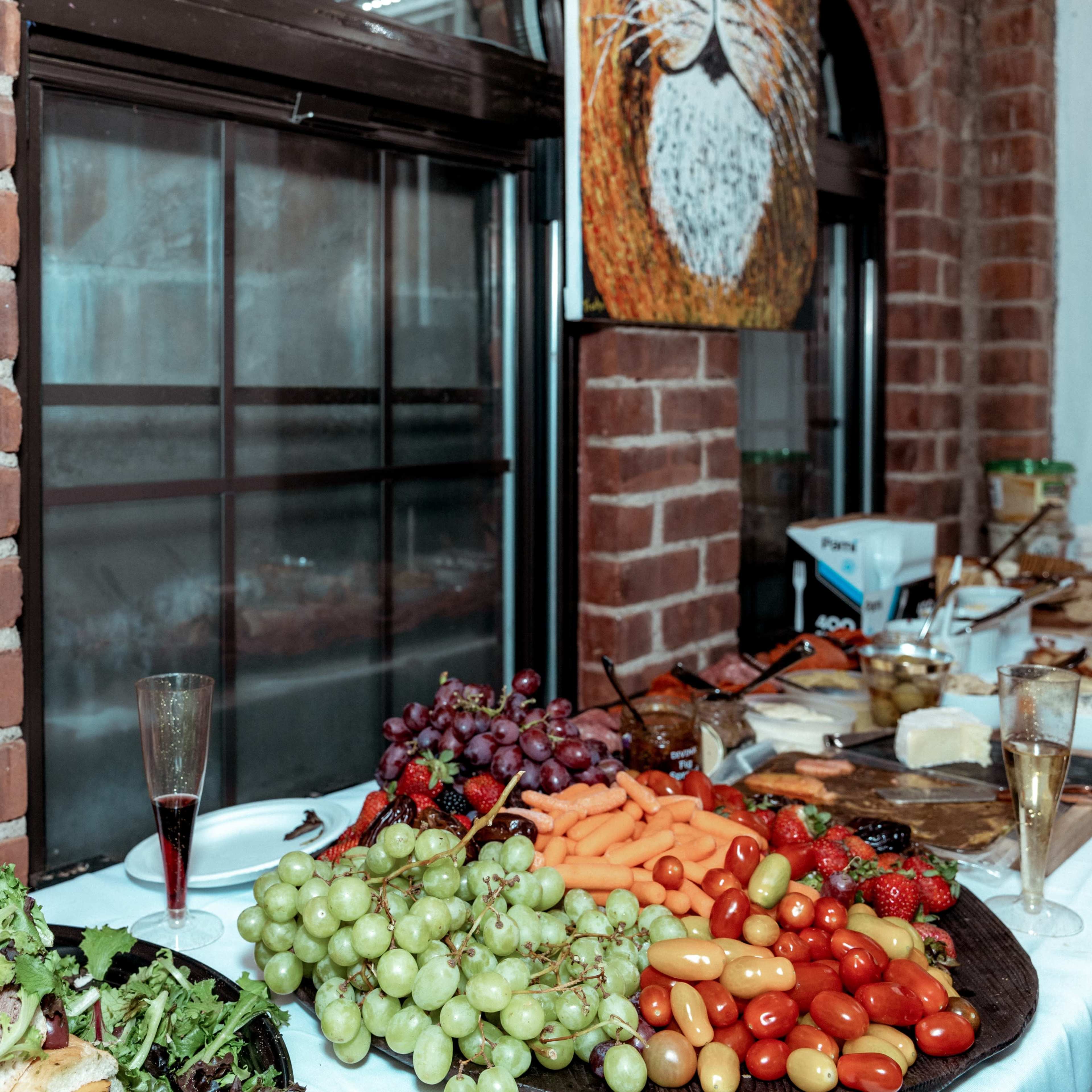A long table displays a variety of food, including sandwiches, grapes, carrots, cherry tomatoes, and strawberries, against a brick wall with an artwork.