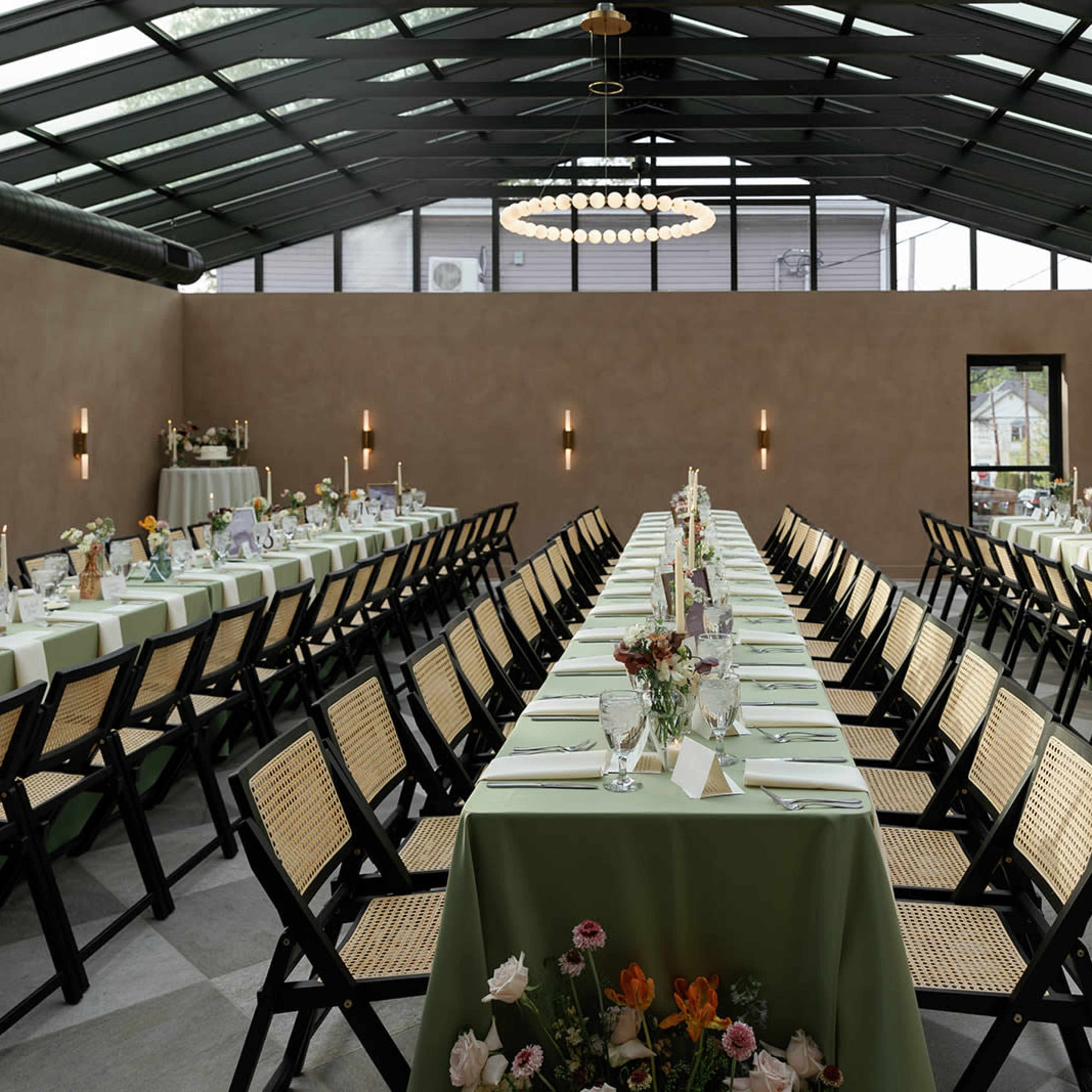 A spacious dining area set with long tables covered in green tablecloths, adorned with floral centerpieces and lit by an overhead chandelier.