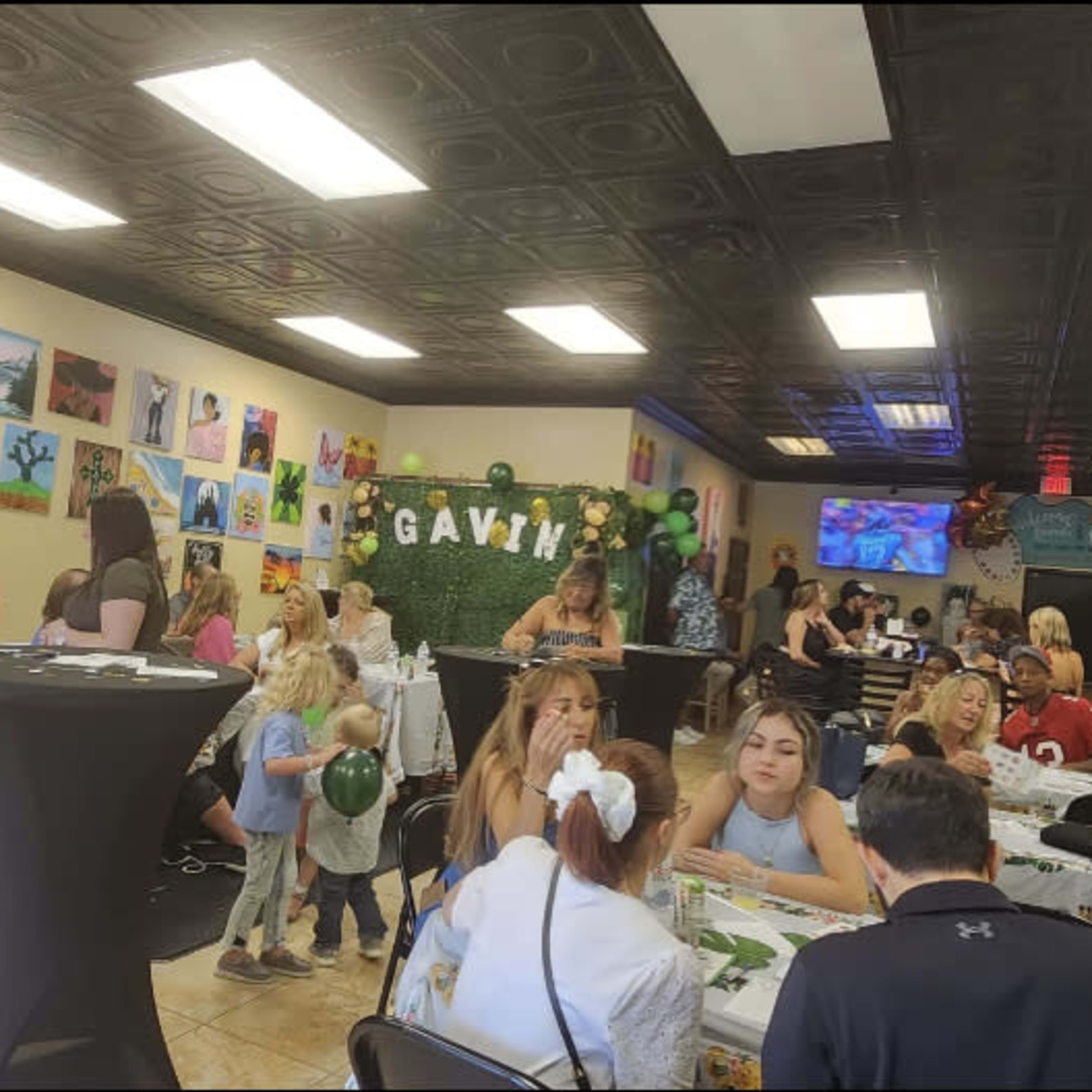 A lively indoor event with guests seated at tables decorated with festive tablecloths, surrounded by artwork on the walls and balloons.