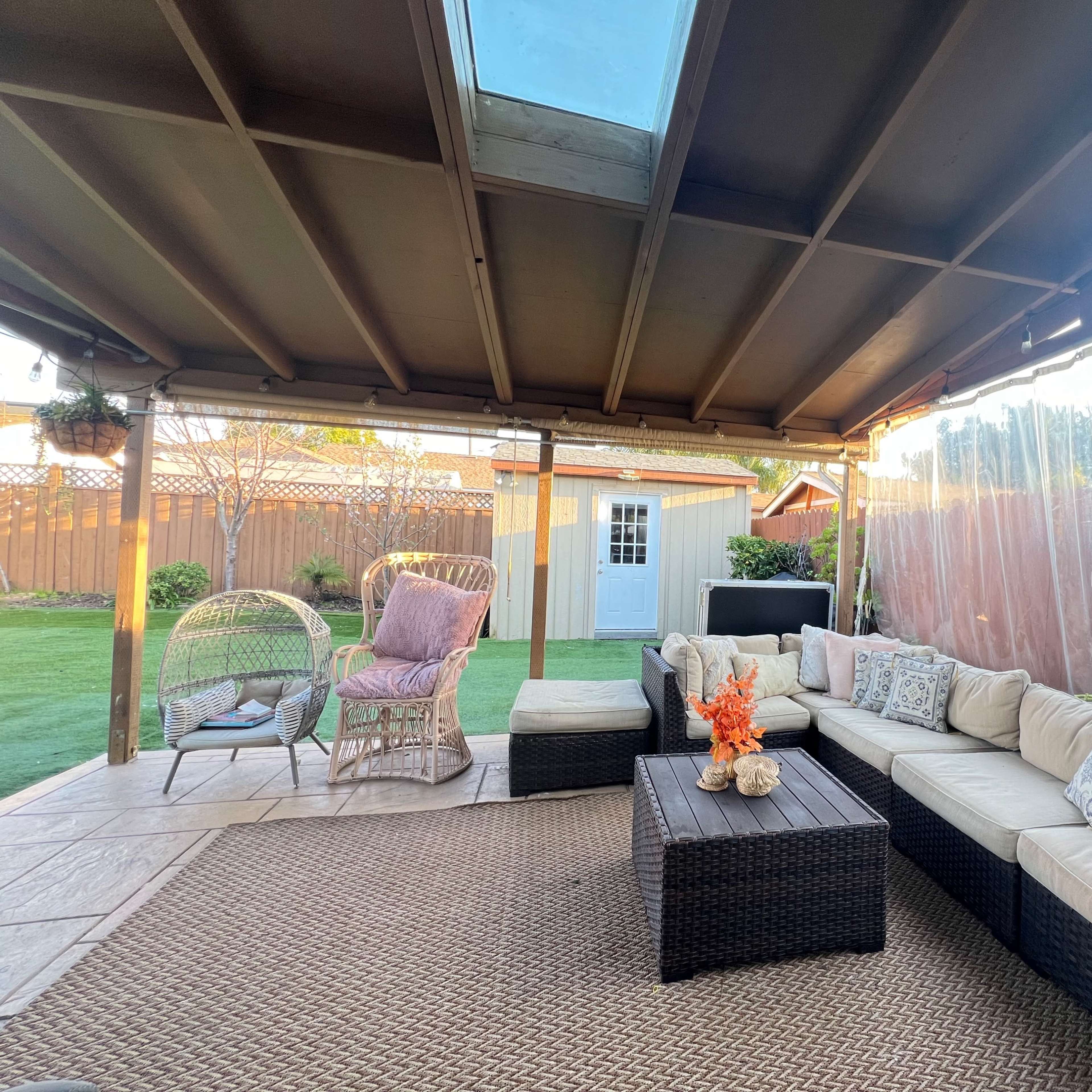 A covered outdoor seating area with a sectional sofa, a coffee table, and decorative elements, adjacent to a grassy backyard with a shed in the background.