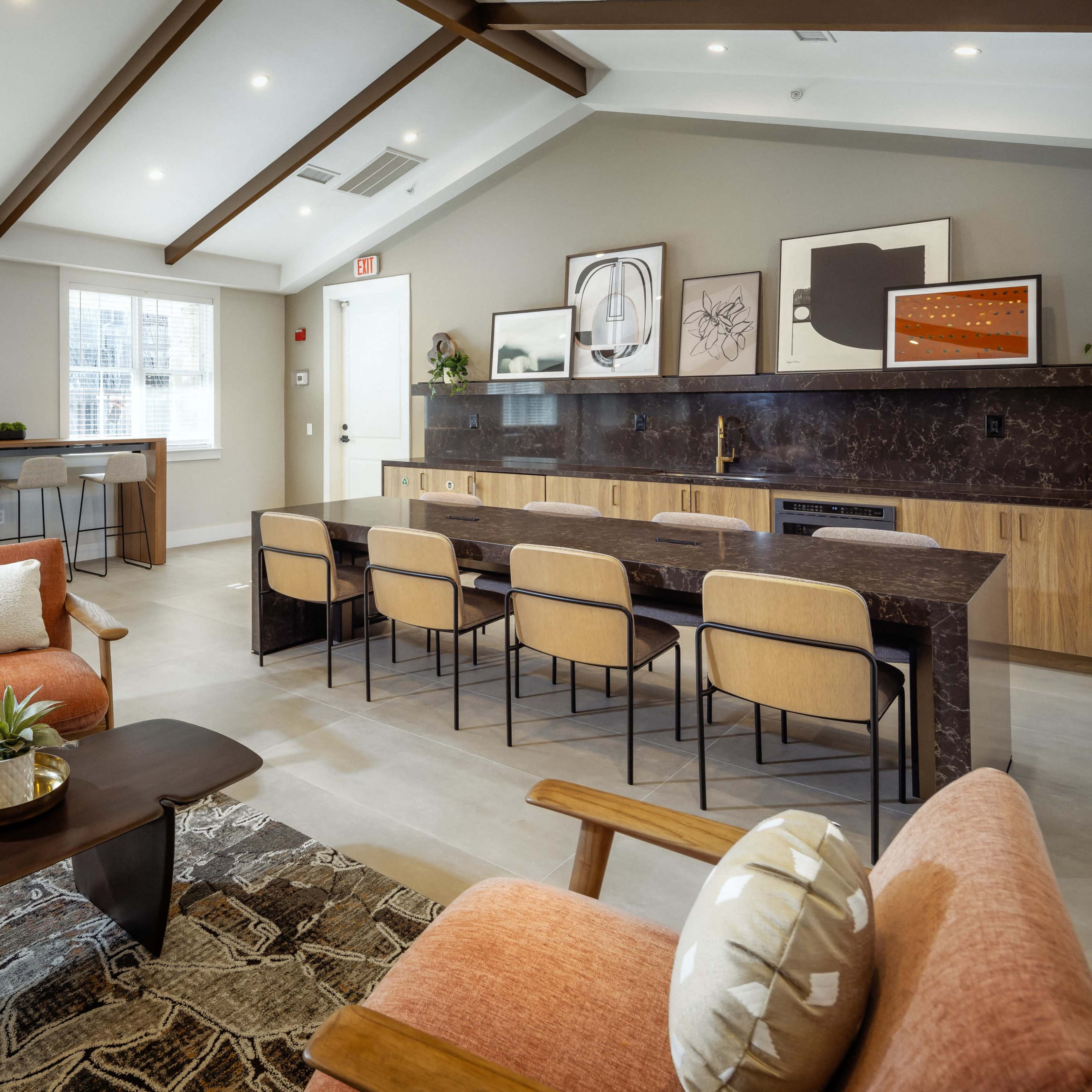 A modern communal space features a dining area with a long marble table, upholstered chairs, and a cozy sitting area with orange sofas and a patterned rug.