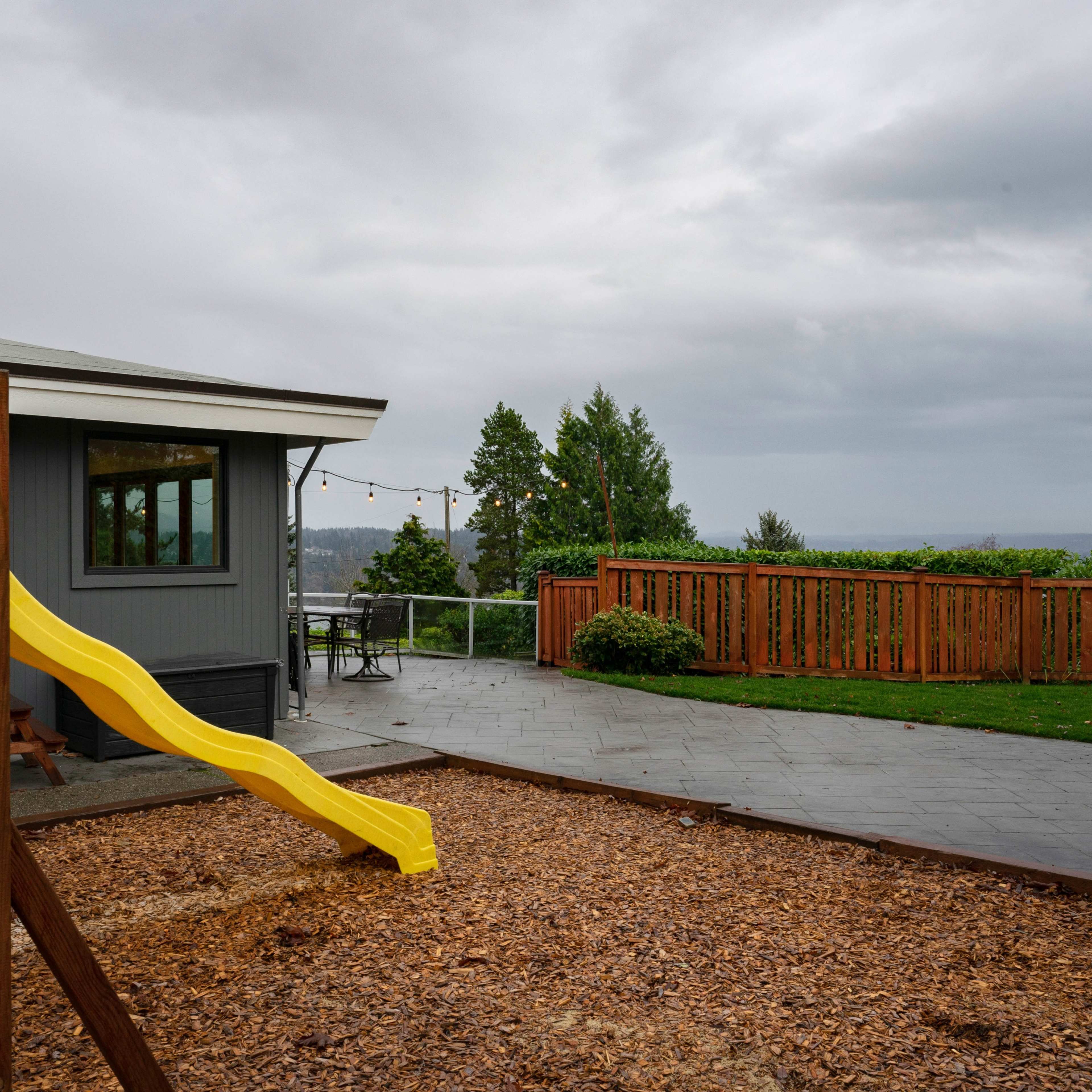 A yellow slide is positioned next to a wooden play structure in a backyard with a patio and a fence under an overcast sky.