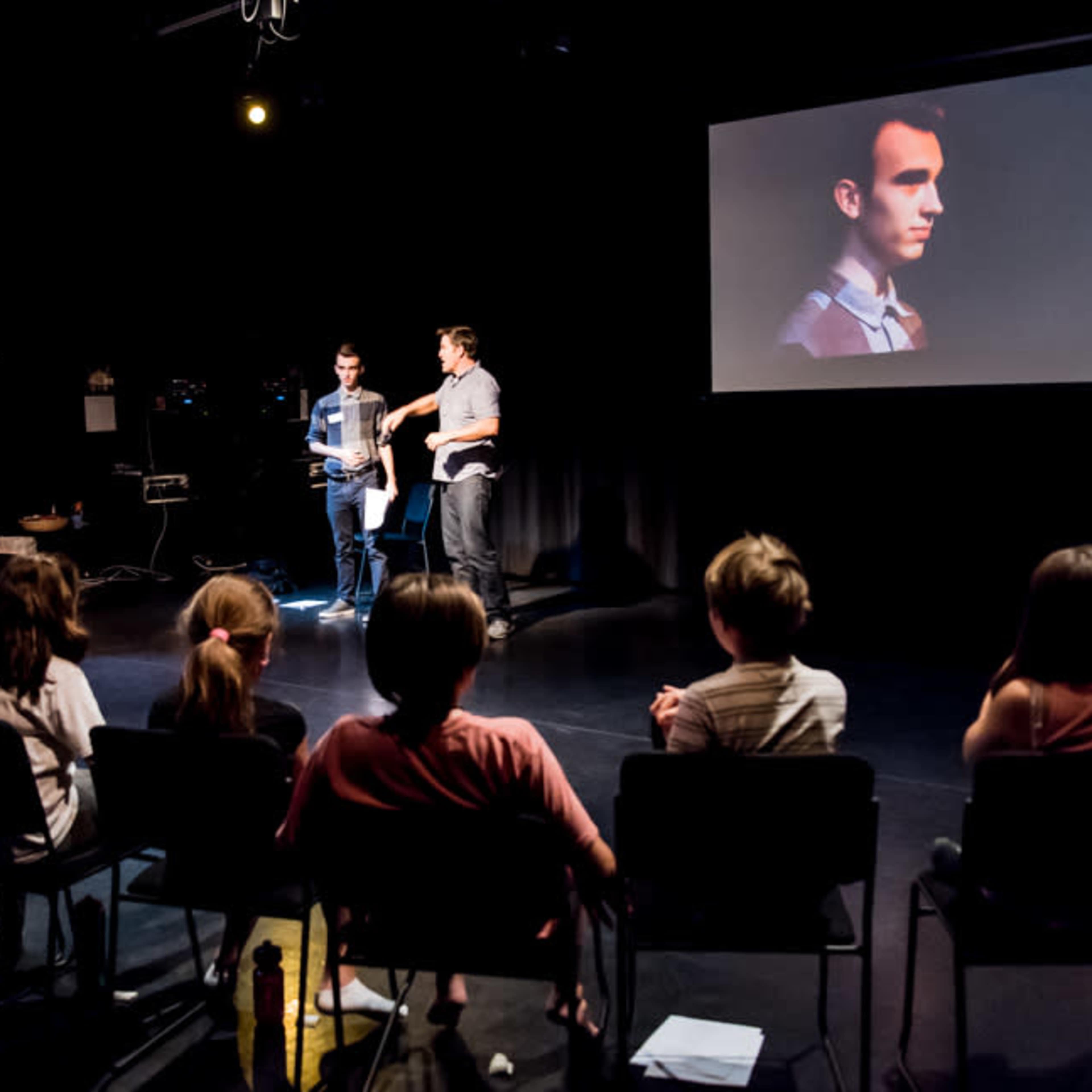 A small group of students watches a presentation in a darkened room with a large screen displaying a man's profile.