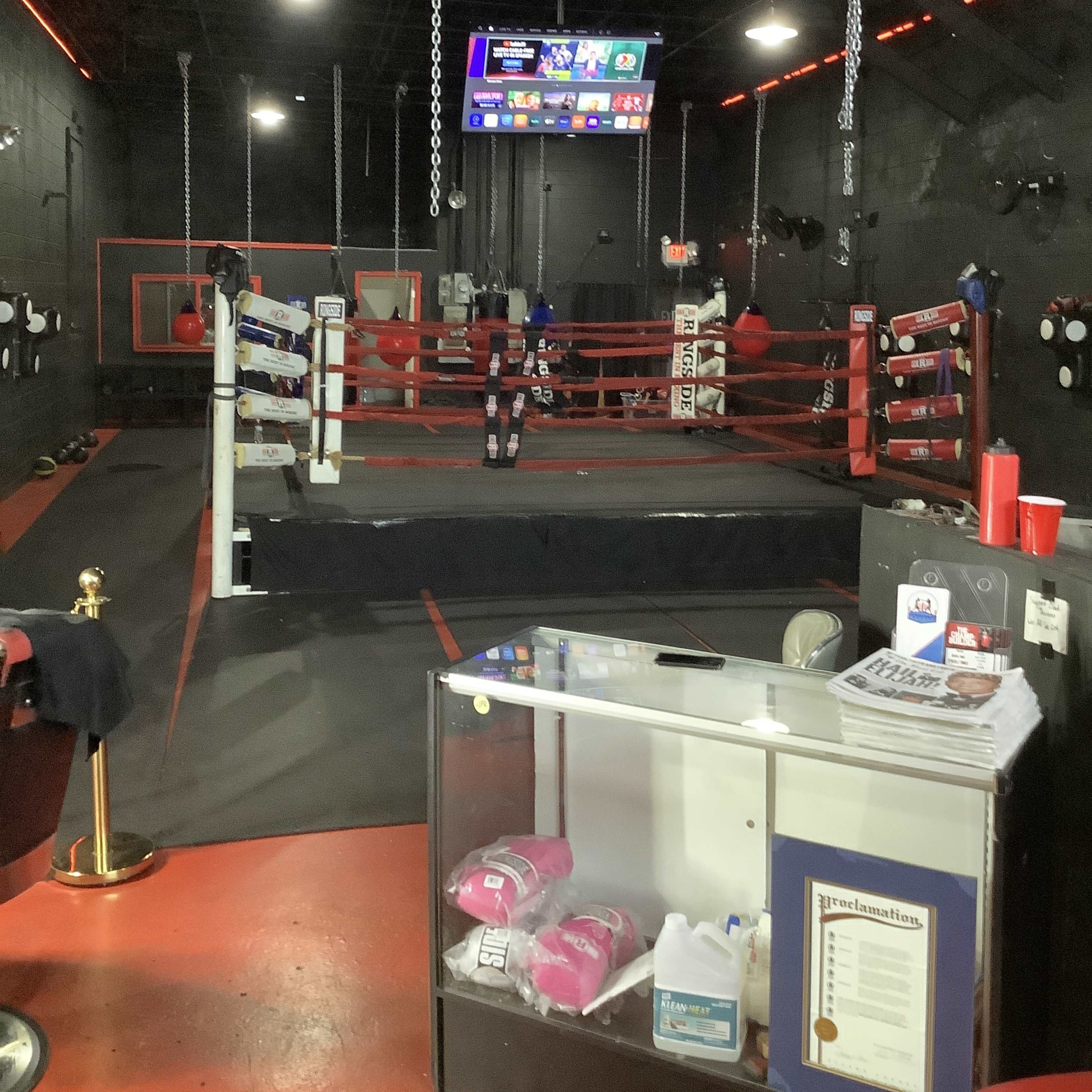 A boxing gym interior with a boxing ring in the center, illuminated by red and white lights, and a reception area with a glass display case in the foreground.