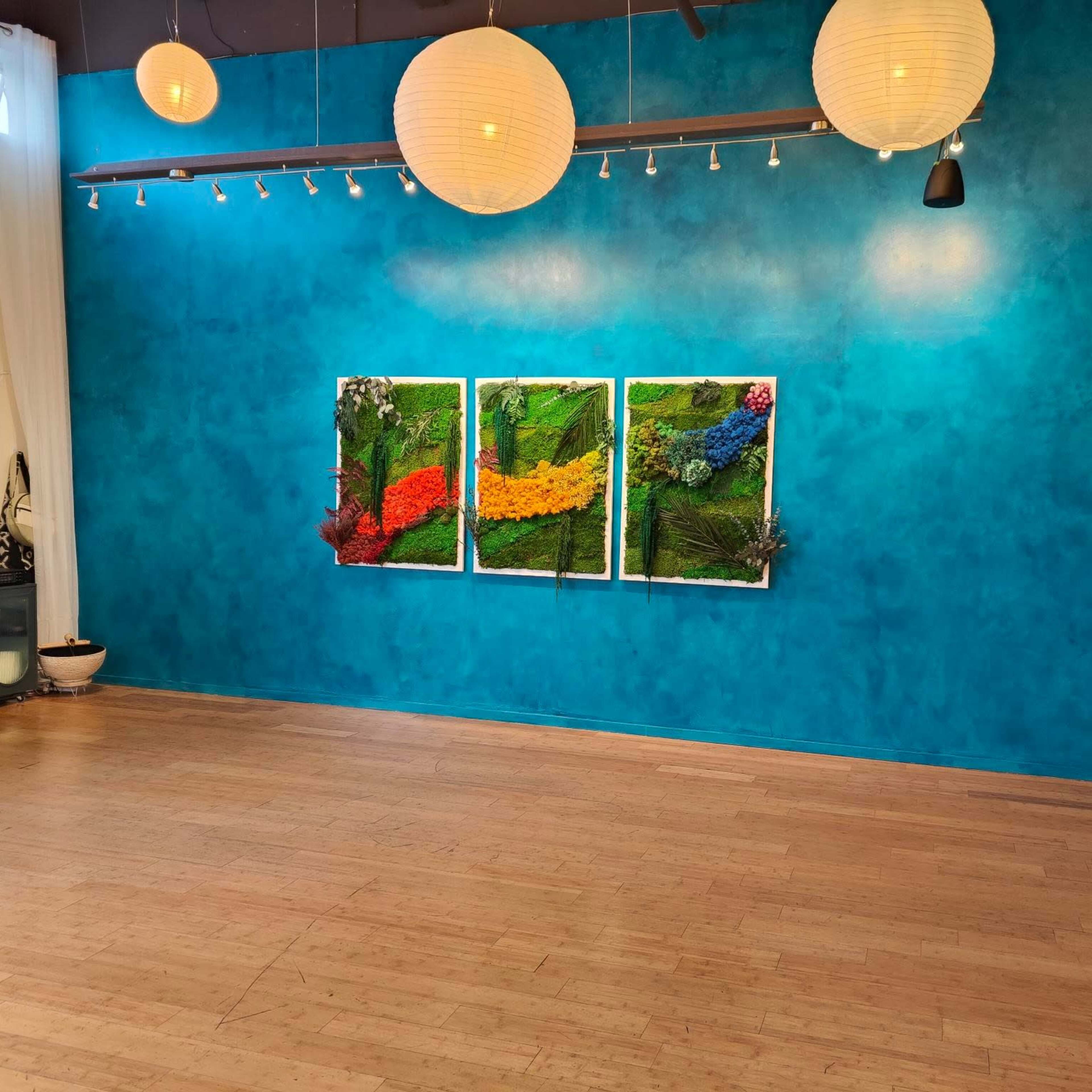 A spacious room with a bright blue wall featuring three framed artworks made of colorful flowers and plants, illuminated by hanging paper lanterns.
