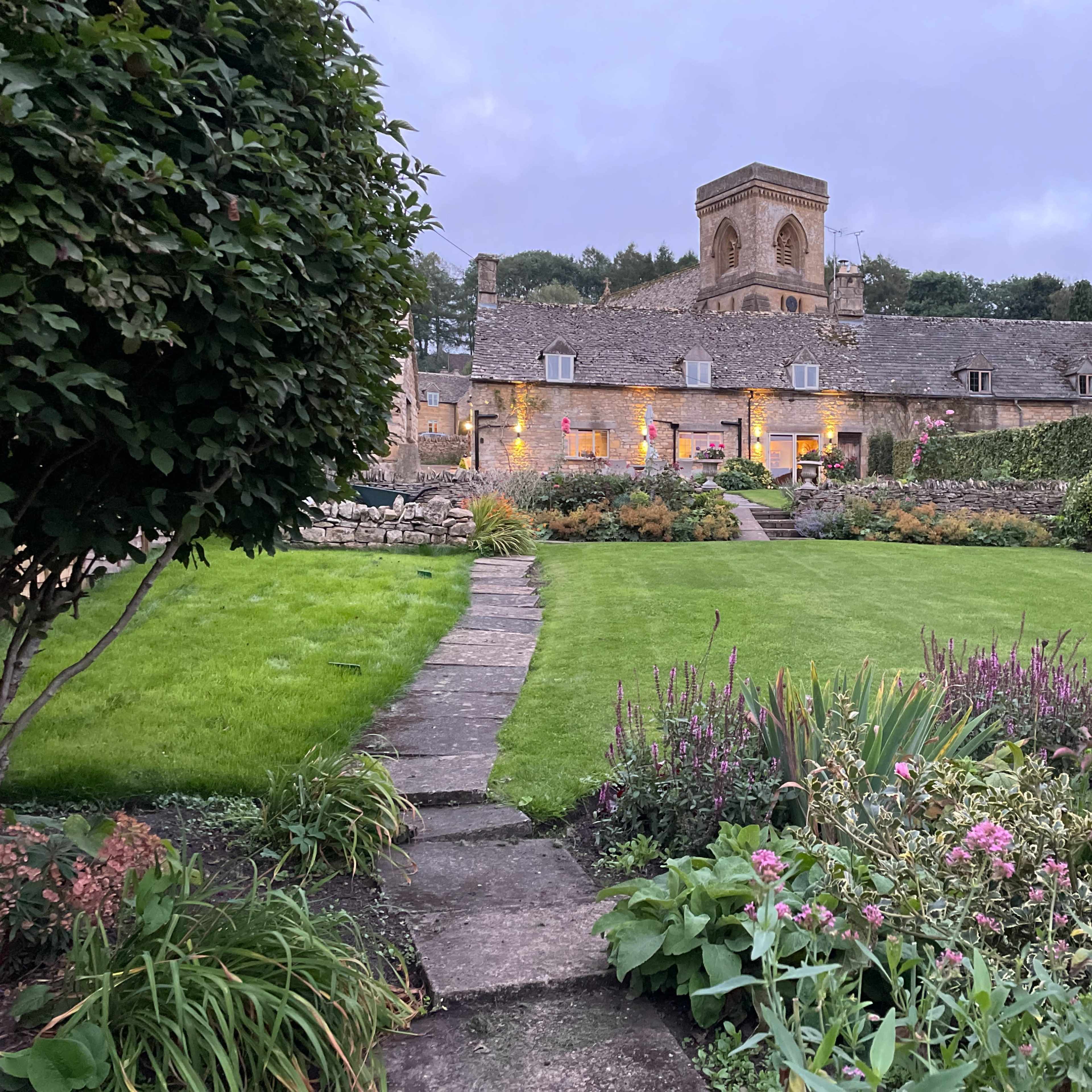 A stone building with a tower is set behind a well-maintained garden featuring a pathway and colorful flowers.