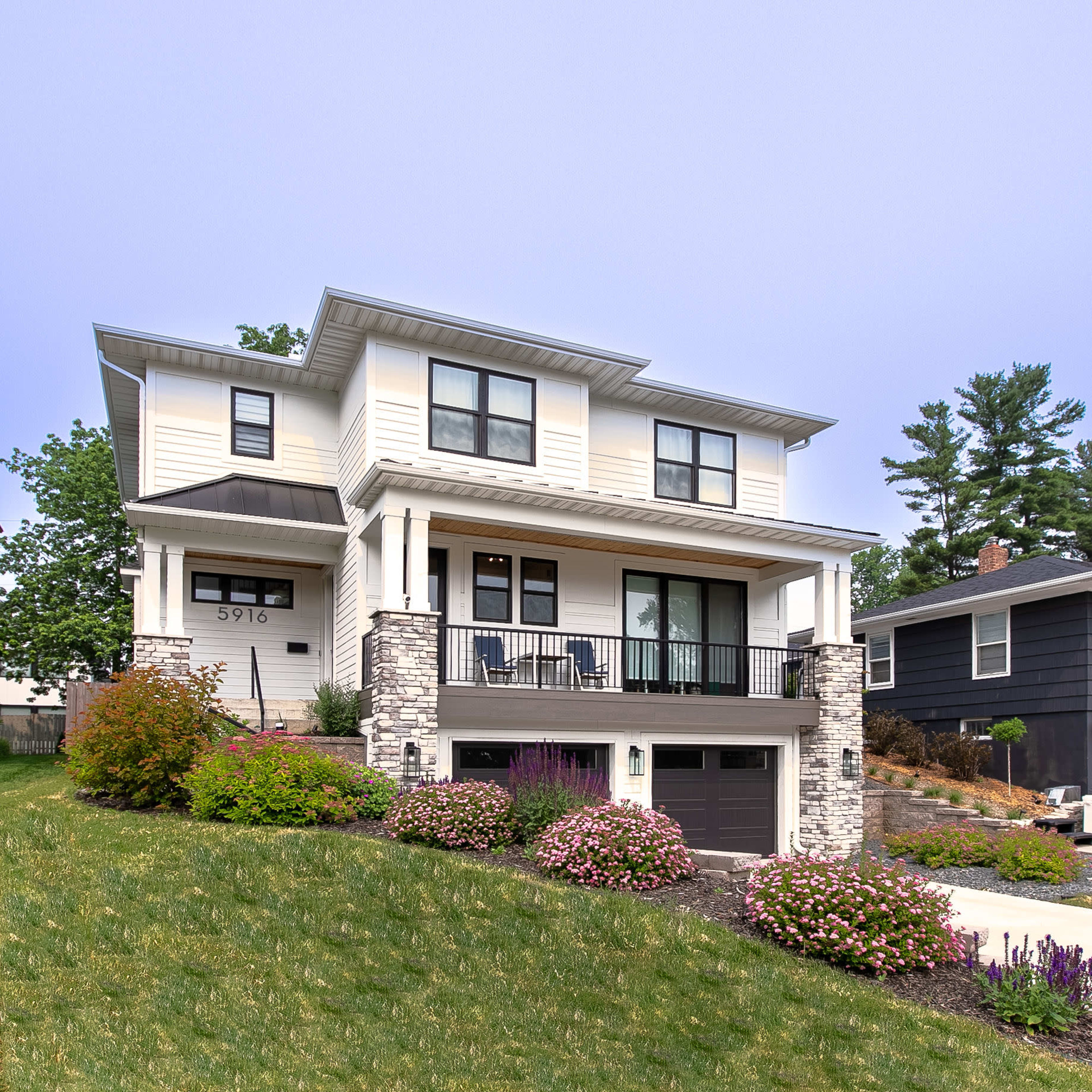 A two-story house with a front porch, stone accents, and landscaping featuring colorful flowers set on a sloping lot.