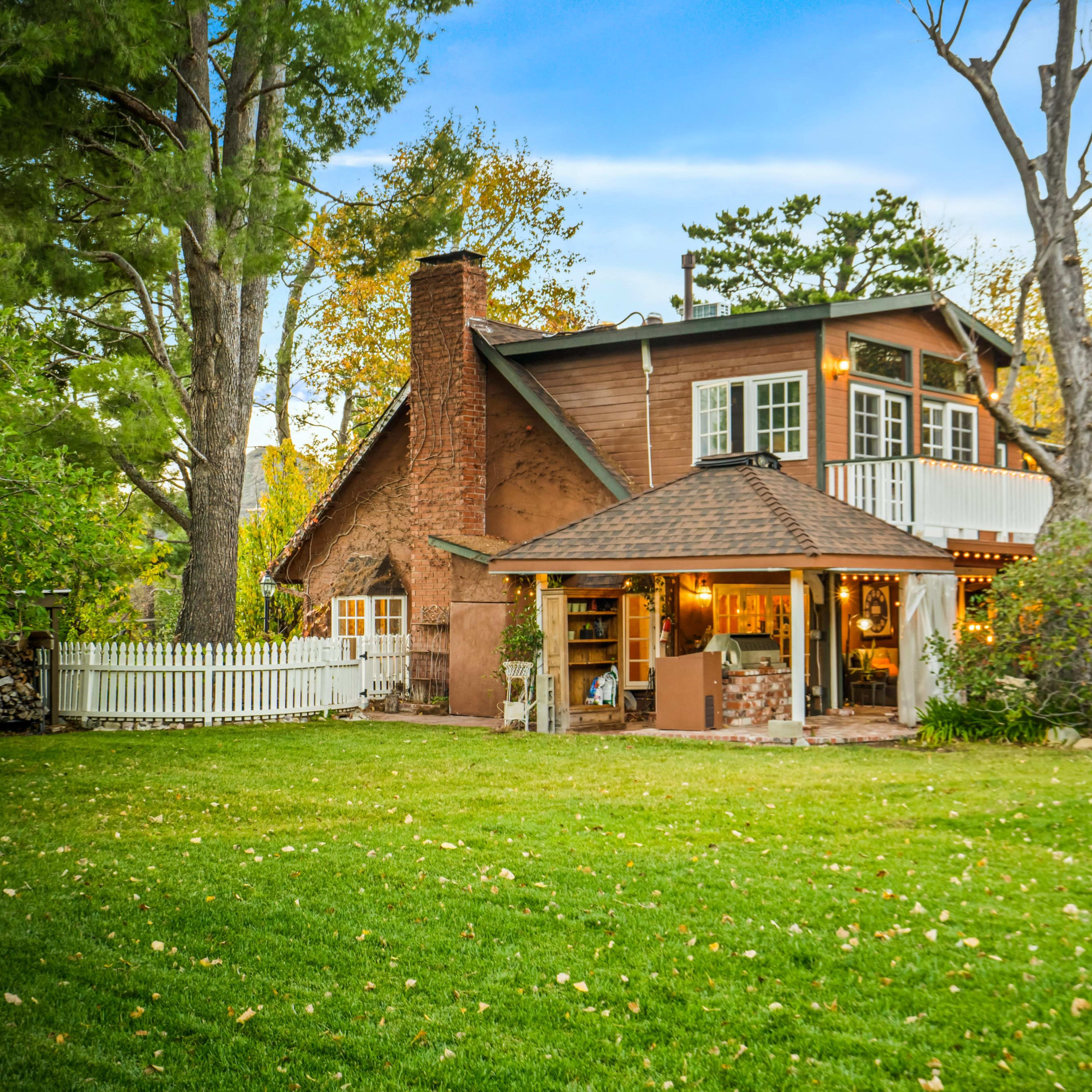 A two-story wooden house with a stone chimney and a covered porch is set amidst a backyard with green grass and trees.