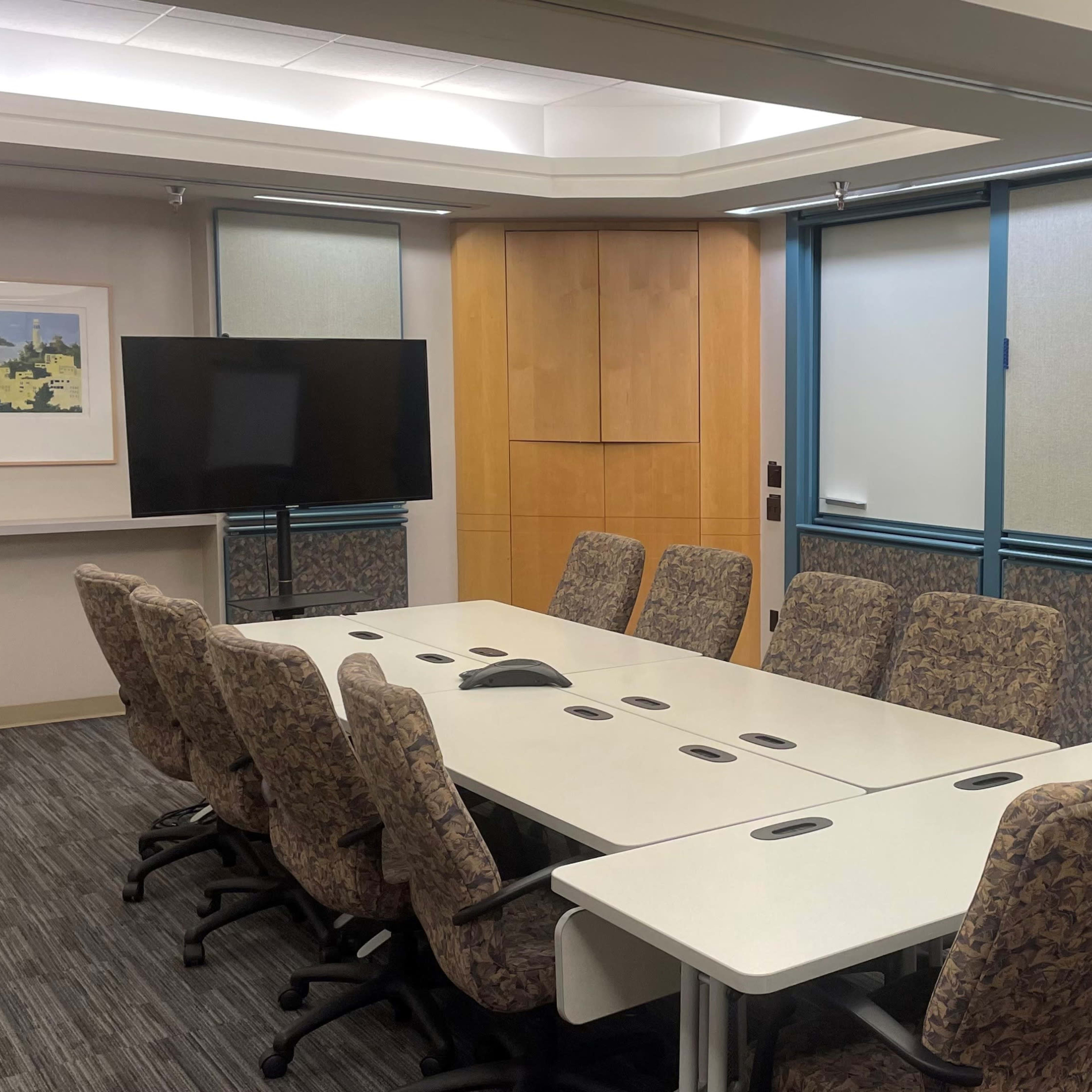 A conference room with a long table, eight chairs, a television screen, and large windows covered by blinds.