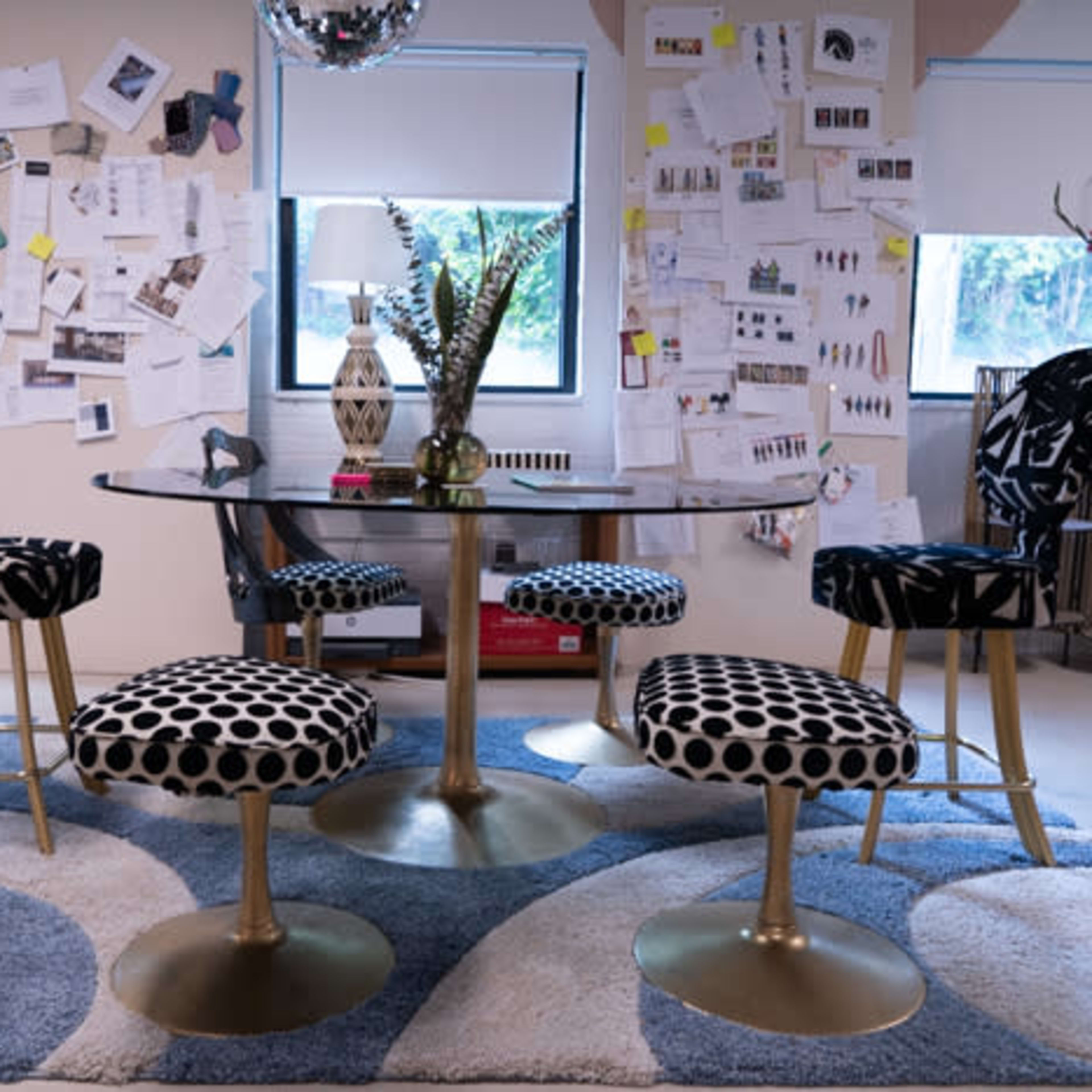 A dining area with a glass table surrounded by upholstered chairs and a patterned rug, against a backdrop of a wall covered with various notes and sketches.