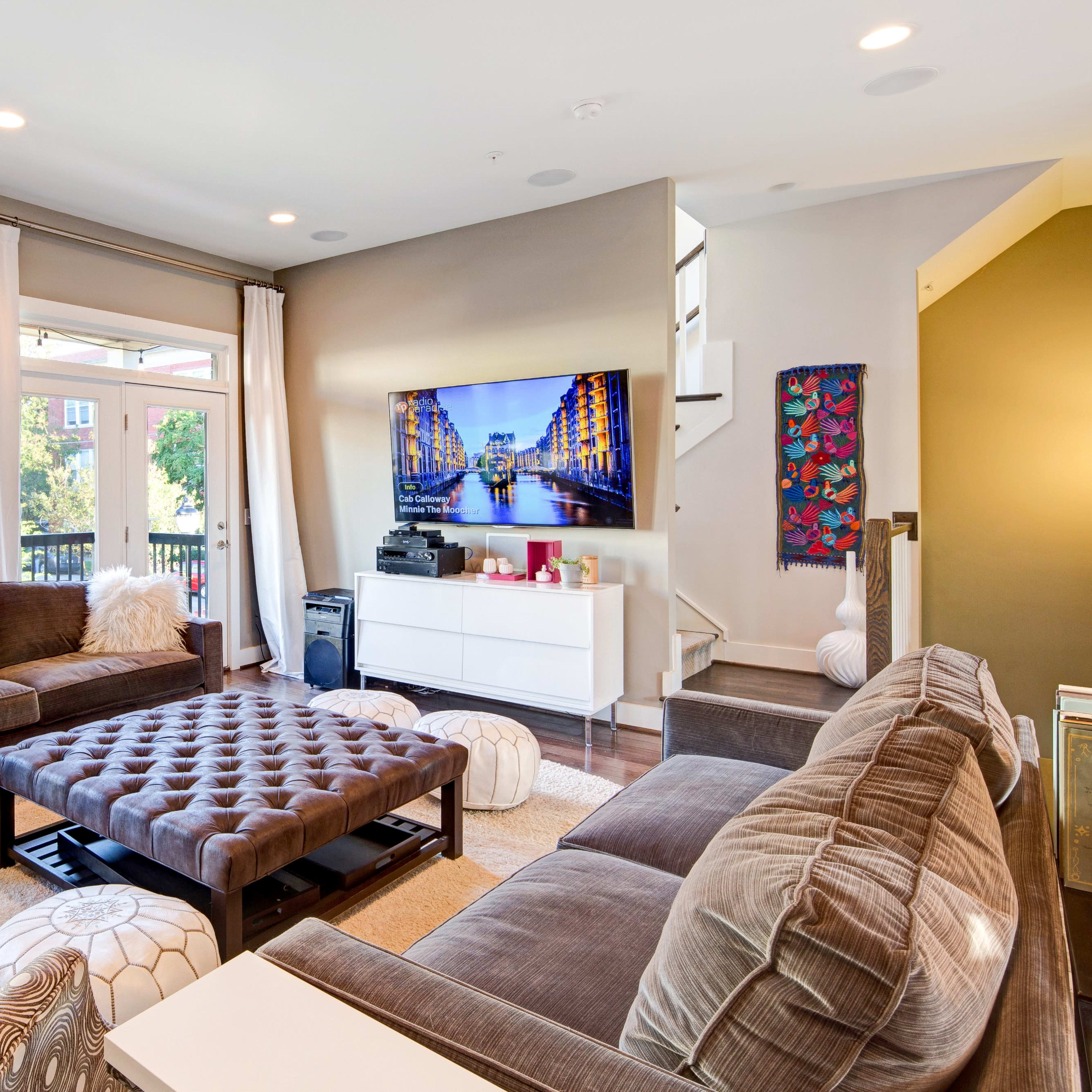 The living room features a sectional sofa, a large coffee table, and a wall-mounted television, with windows providing natural light and a view of the outdoors.