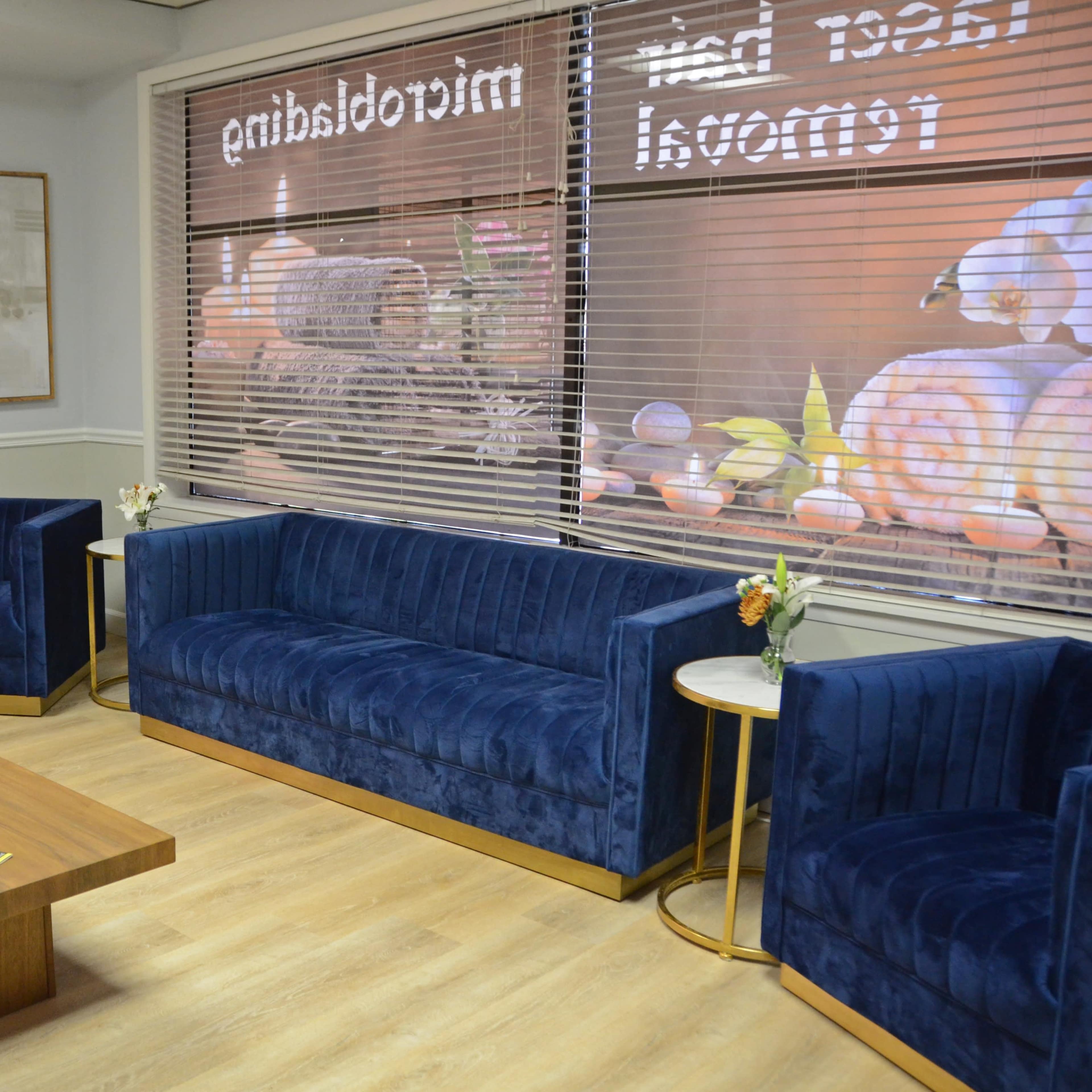 A waiting area with blue velvet chairs and a sofa, a wooden coffee table, and large windows featuring a decorative display.