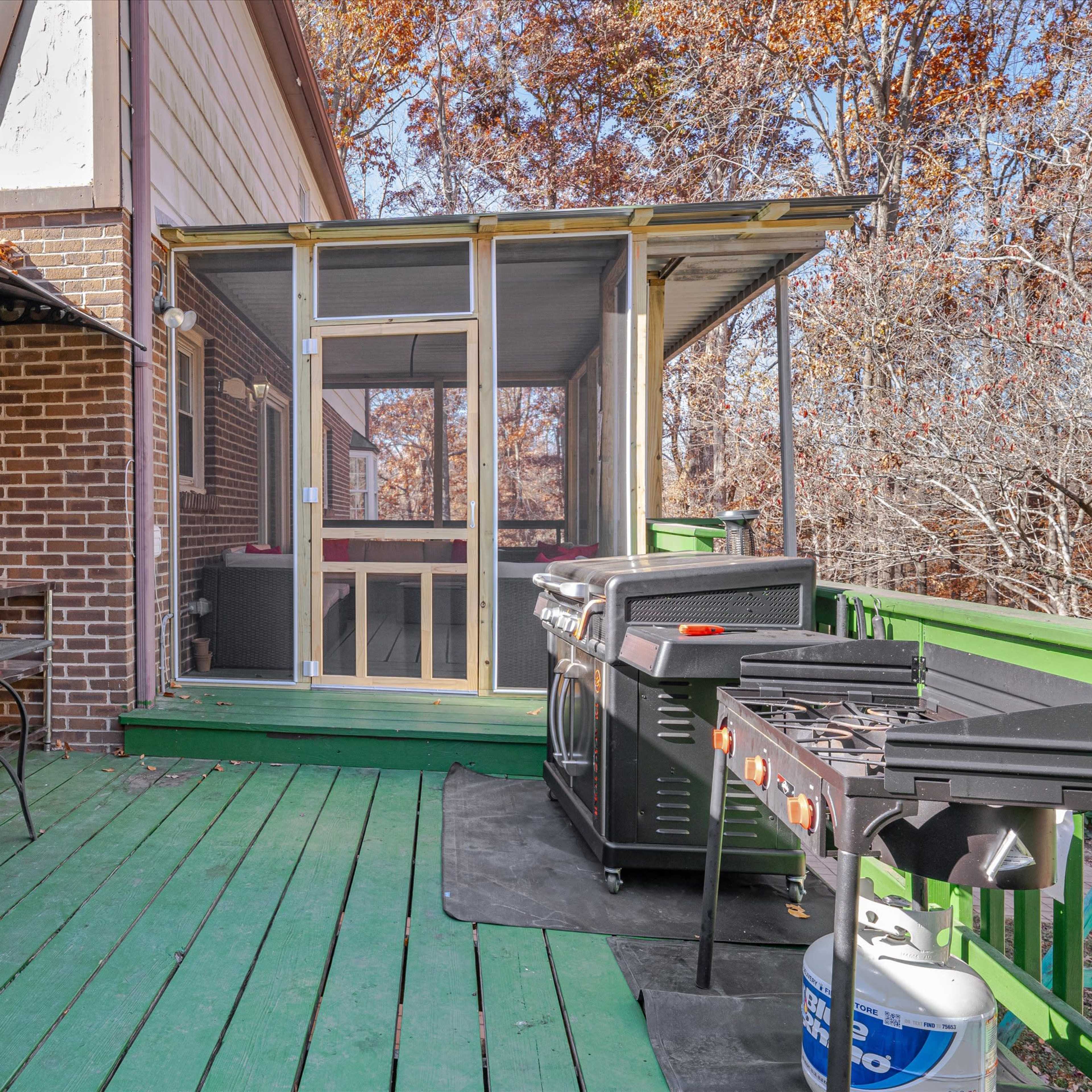 An outdoor space with a green wooden deck, a brick wall, a screened patio entrance, and a barbecue grill setup.