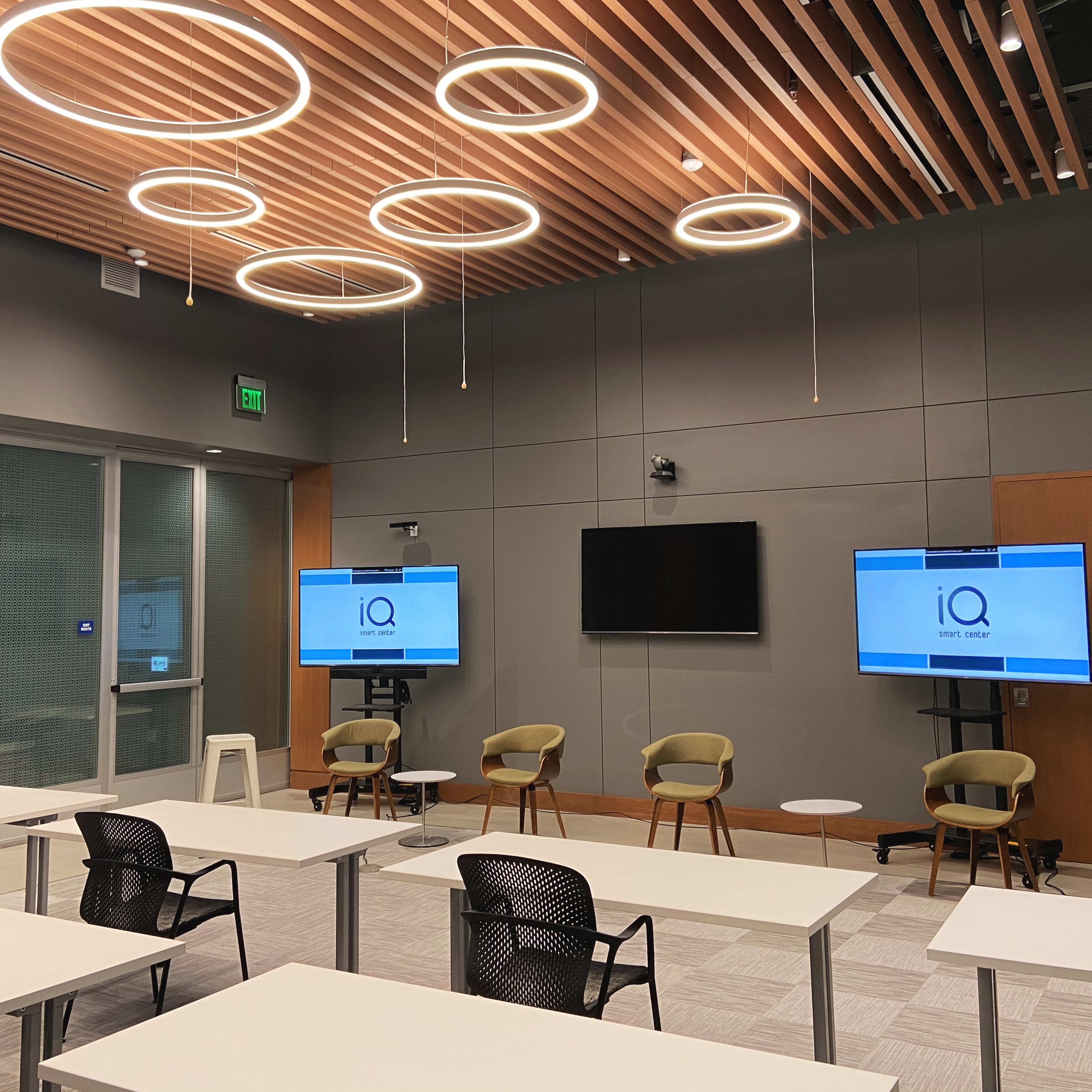 A modern conference room features multiple circular lights, two screens displaying the logo "iQ," and several tables and chairs arranged for a meeting.