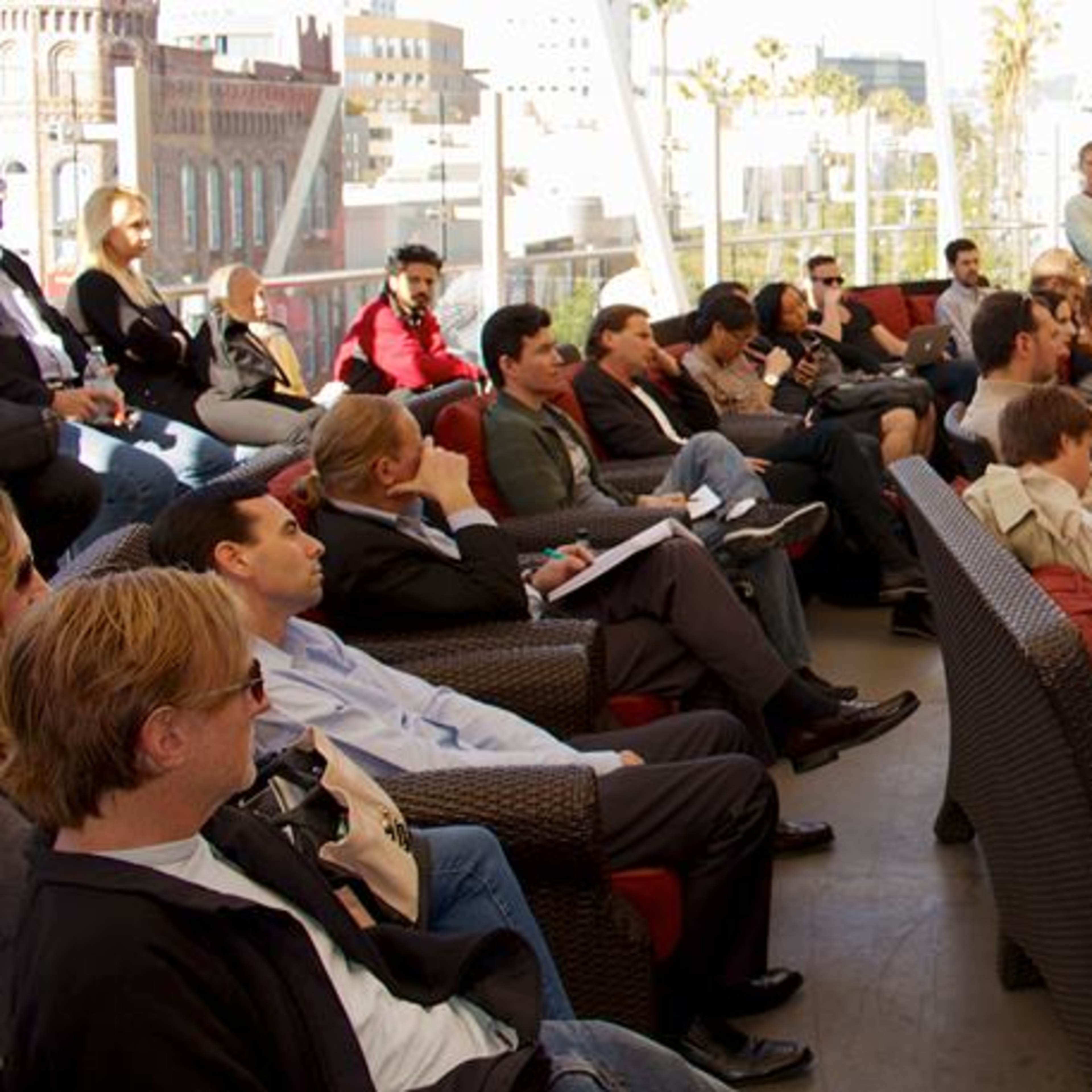 A group of people sits in a tiered outdoor space, attentively watching two speakers engaged in conversation on a stage.