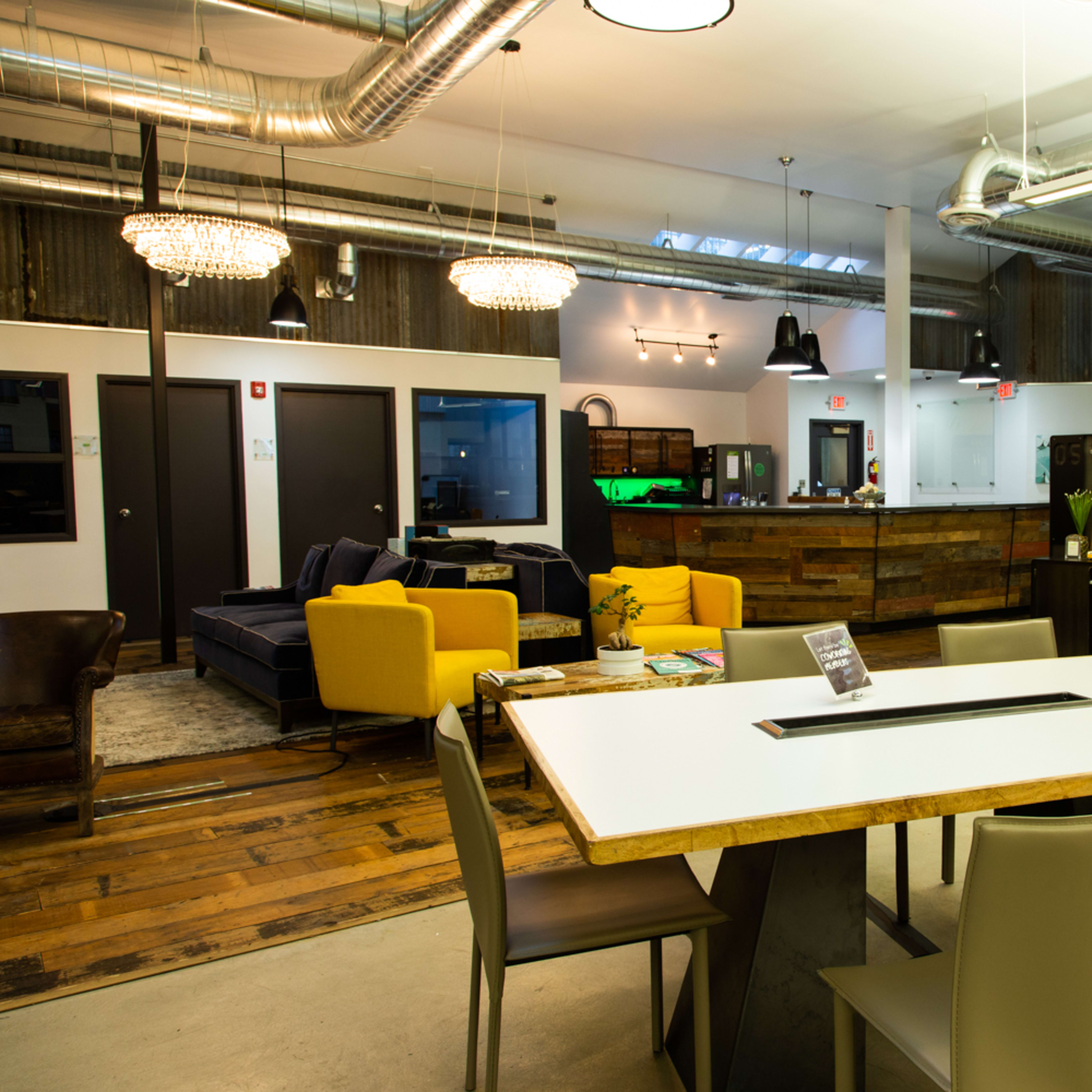 A modern coworking space featuring a communal table, chairs, a lounge area, and an open reception area with various seating arrangements.
