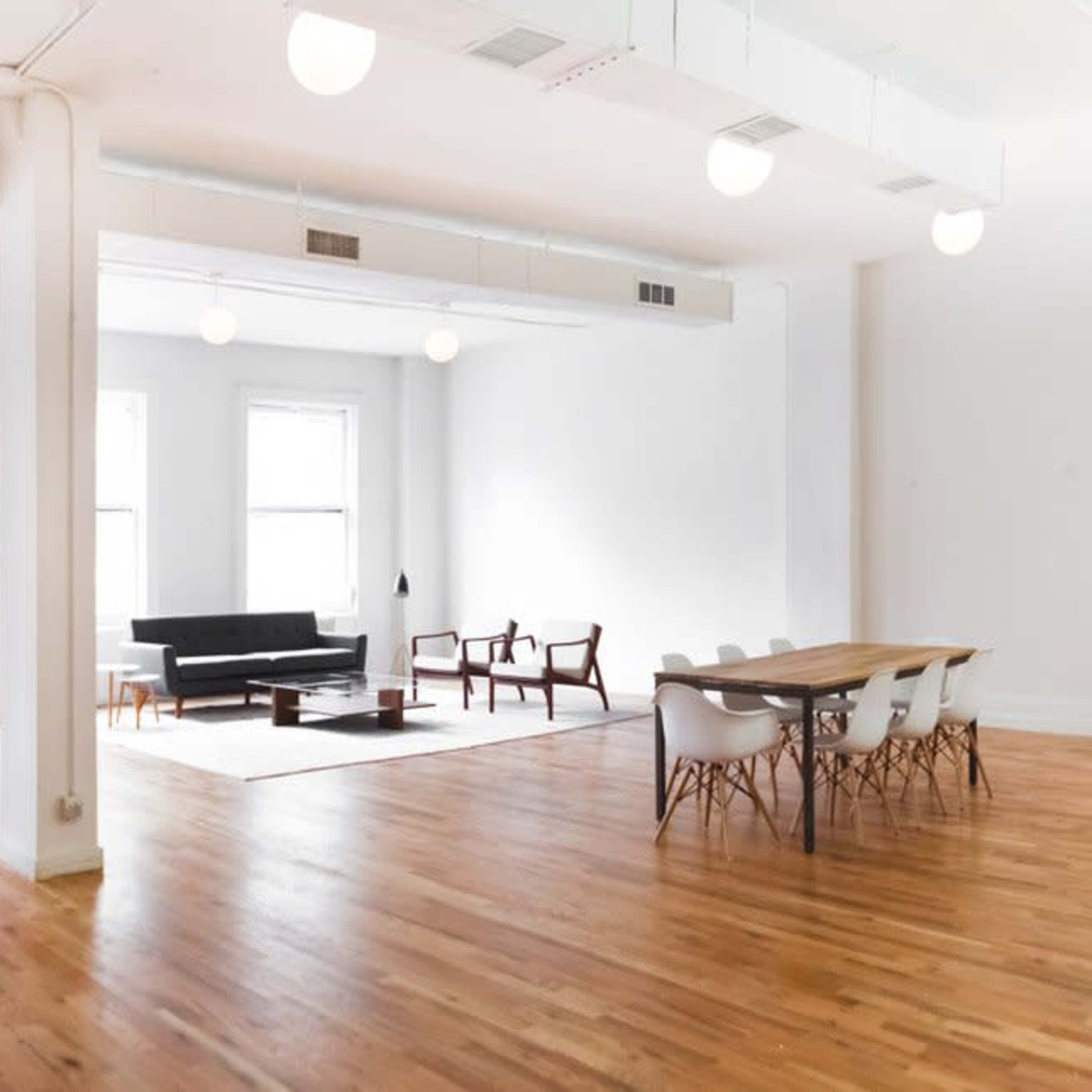 A spacious, minimalistic room featuring large windows, hardwood flooring, and various modern furniture pieces including a sofa, chairs, and a dining table.