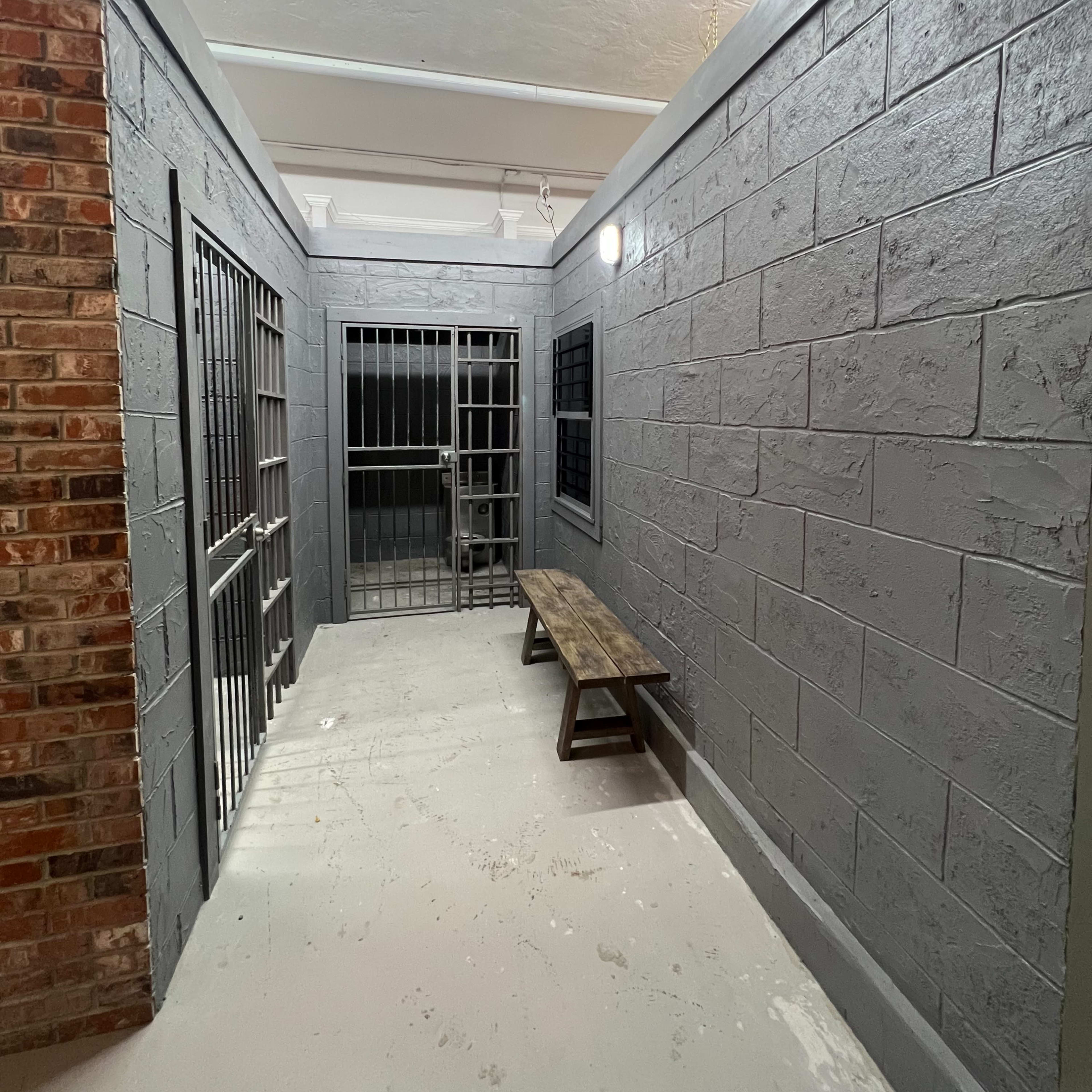 A hallway in a facility featuring gray walls, metal bars on two doors, and a wooden bench against one side.