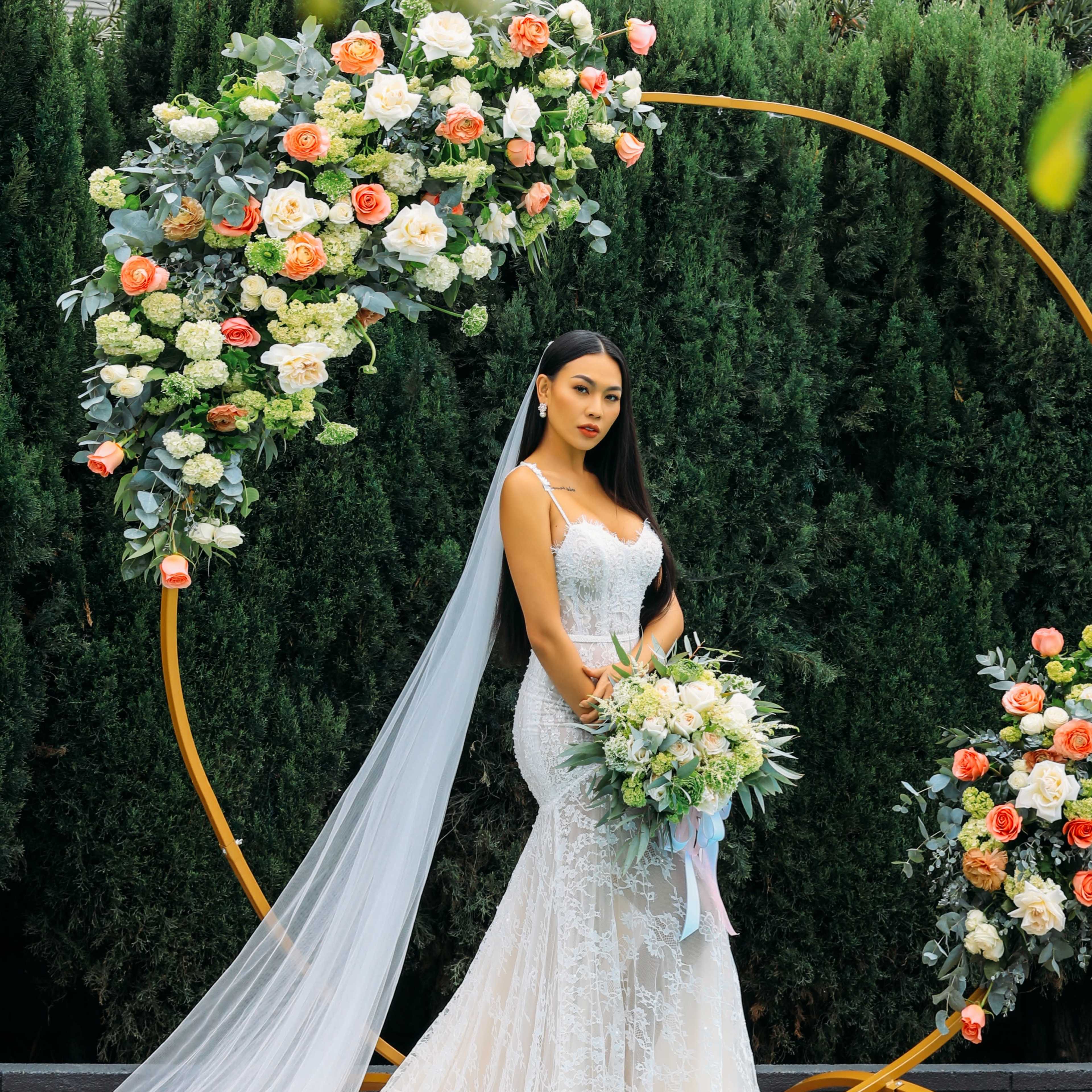 A bride in a fitted lace gown poses with a bouquet in front of a floral arch adorned with roses and greenery.